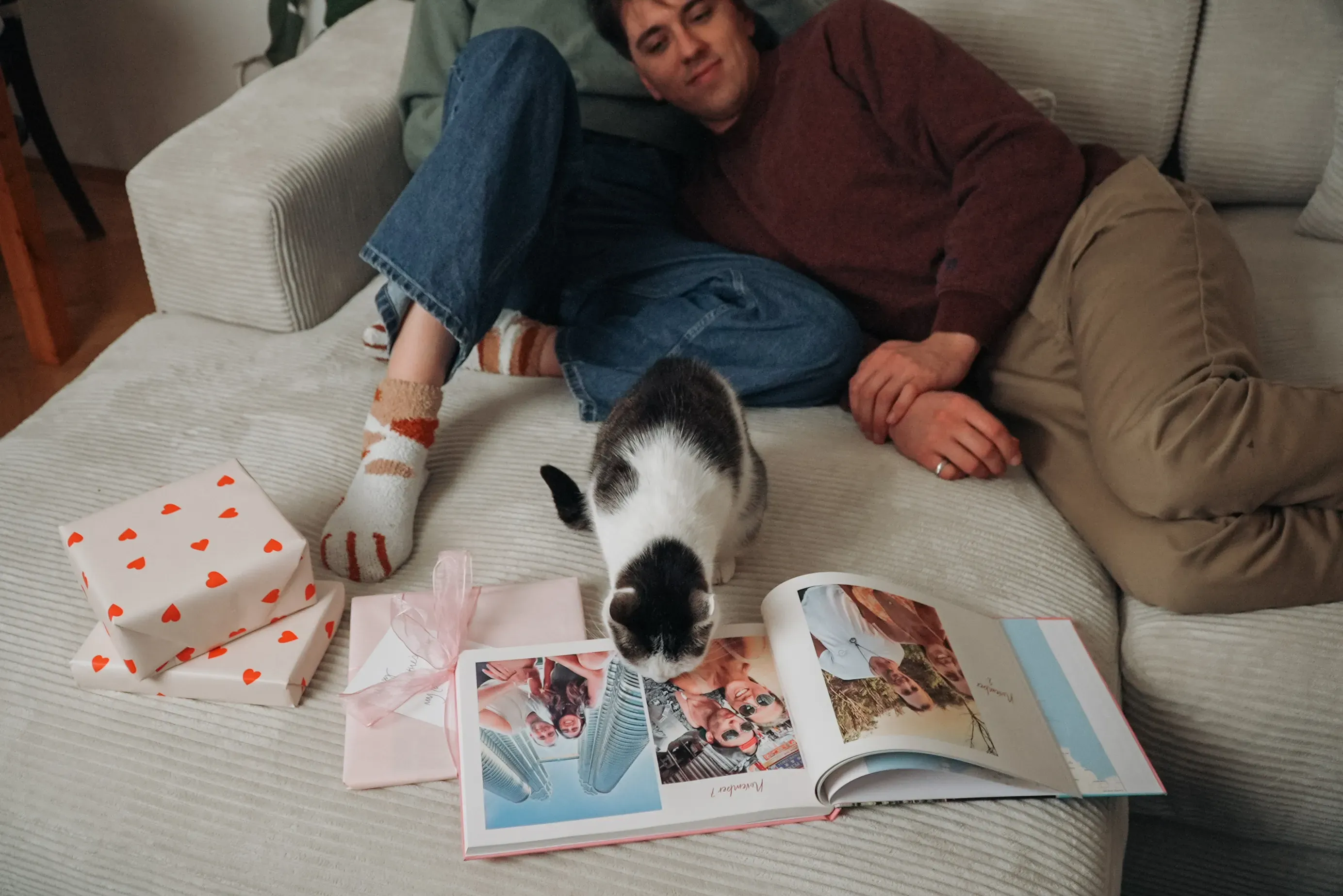 A couple relaxes on a couch with a cat curiously looking at an open photo album, surrounded by wrapped gifts.