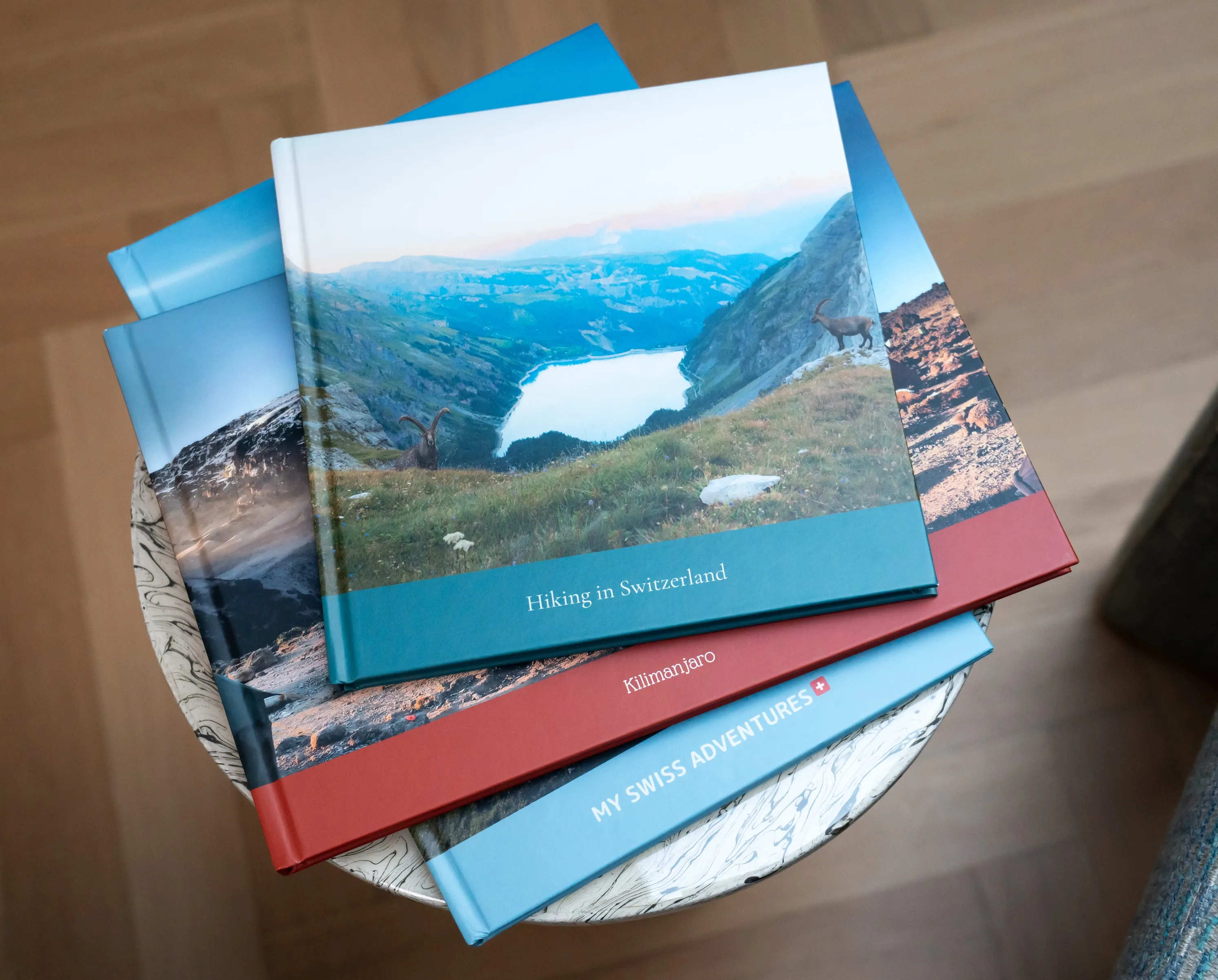 Stack of travel photo books on a round table. Top book shows a mountain landscape with the title "Hiking in Switzerland."