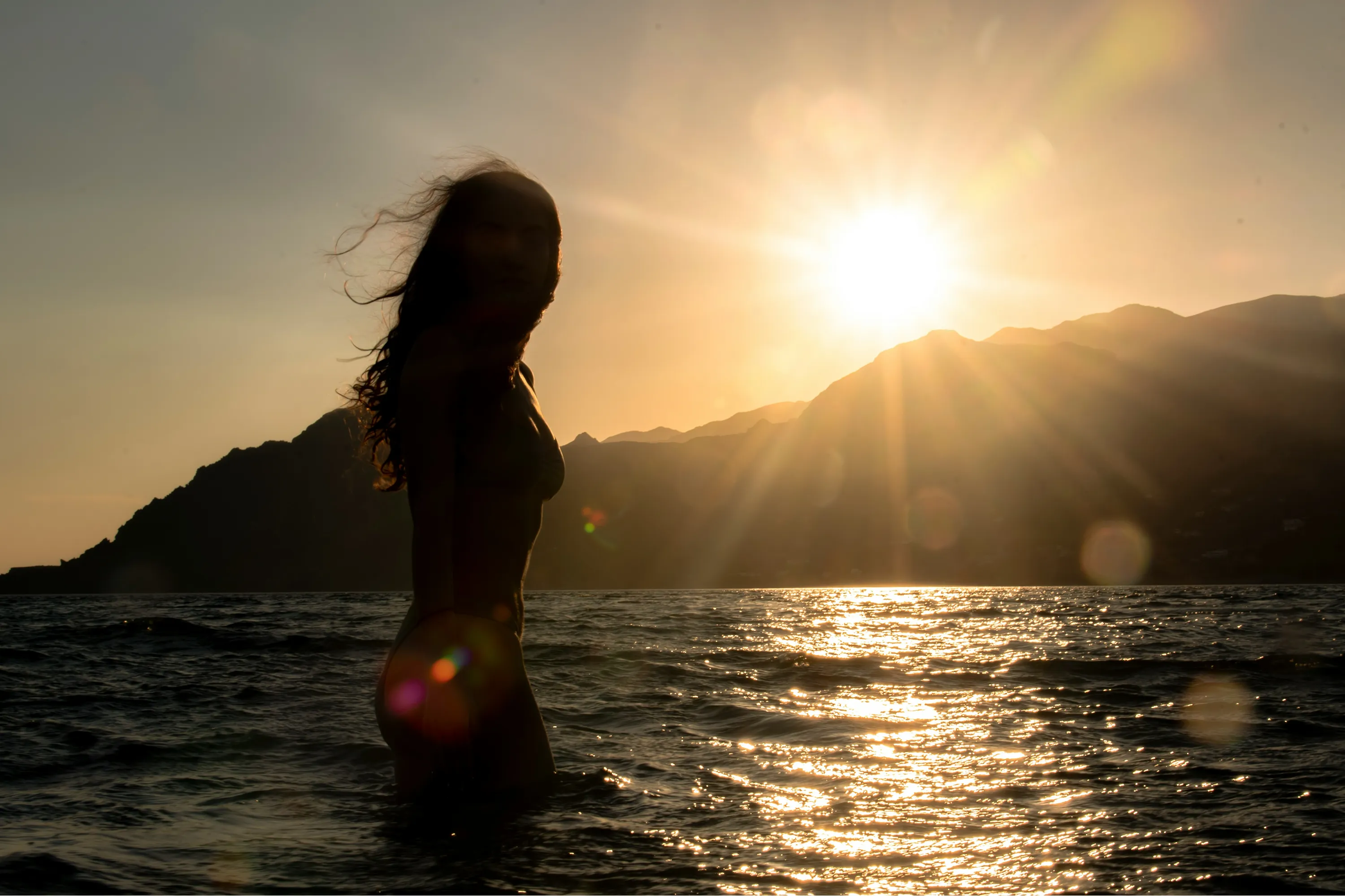 Silhouette d'une personne debout dans l'eau au coucher du soleil, avec des montagnes en arrière-plan et les reflets du soleil sur la mer.