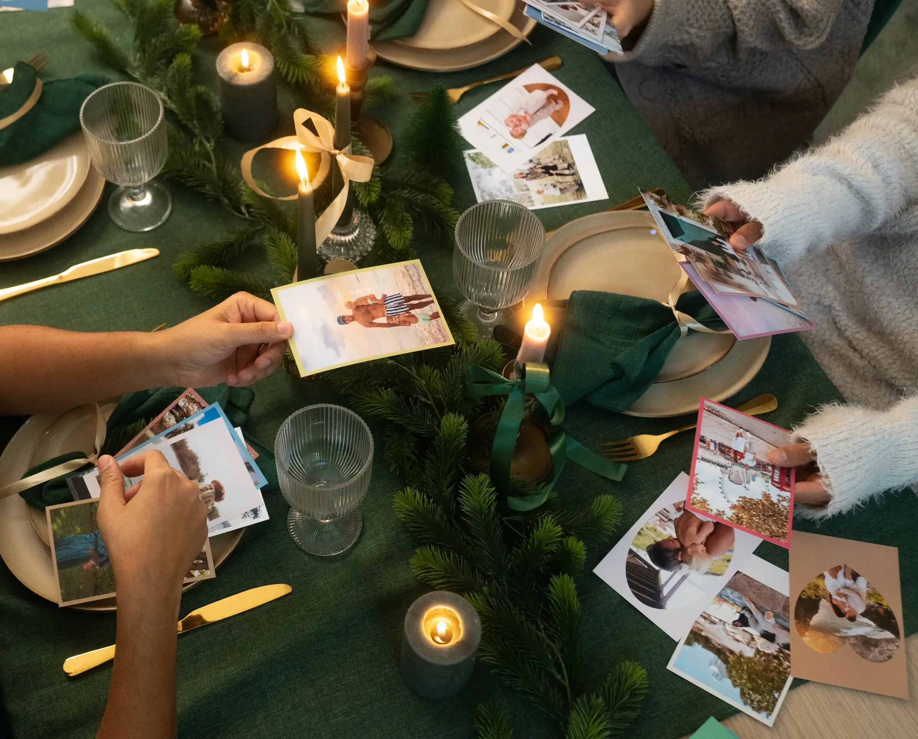 People sharing photos around a candlelit table with green decor, gold cutlery, and pine branches, creating a warm, festive atmosphere.