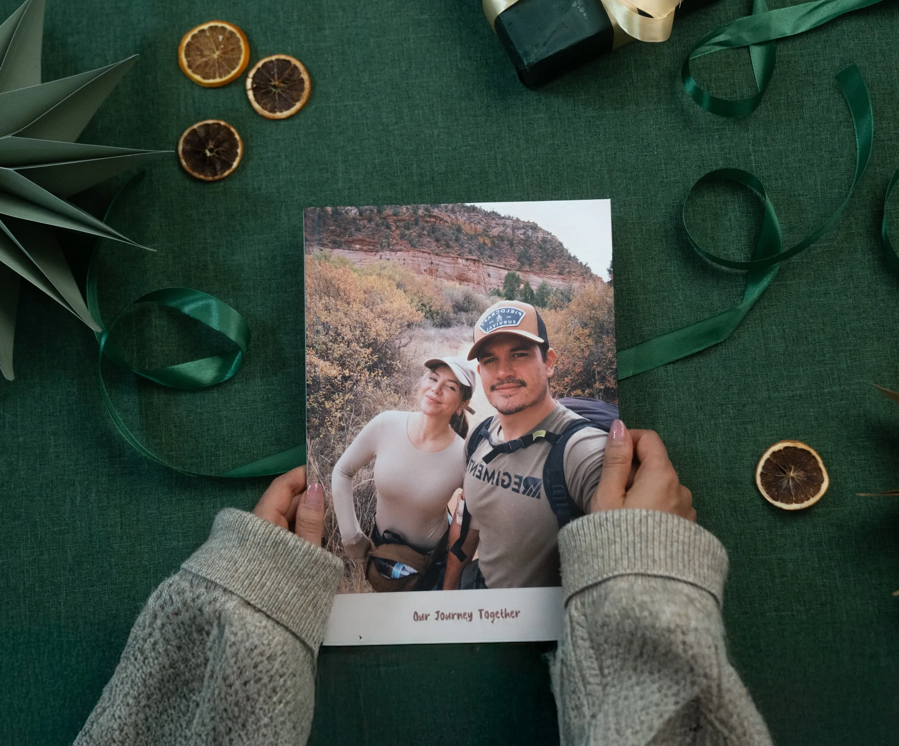 Hands holding a photo book titled "Our Journey Together" with a cover image of a couple hiking, surrounded by festive decorations on a green surface.