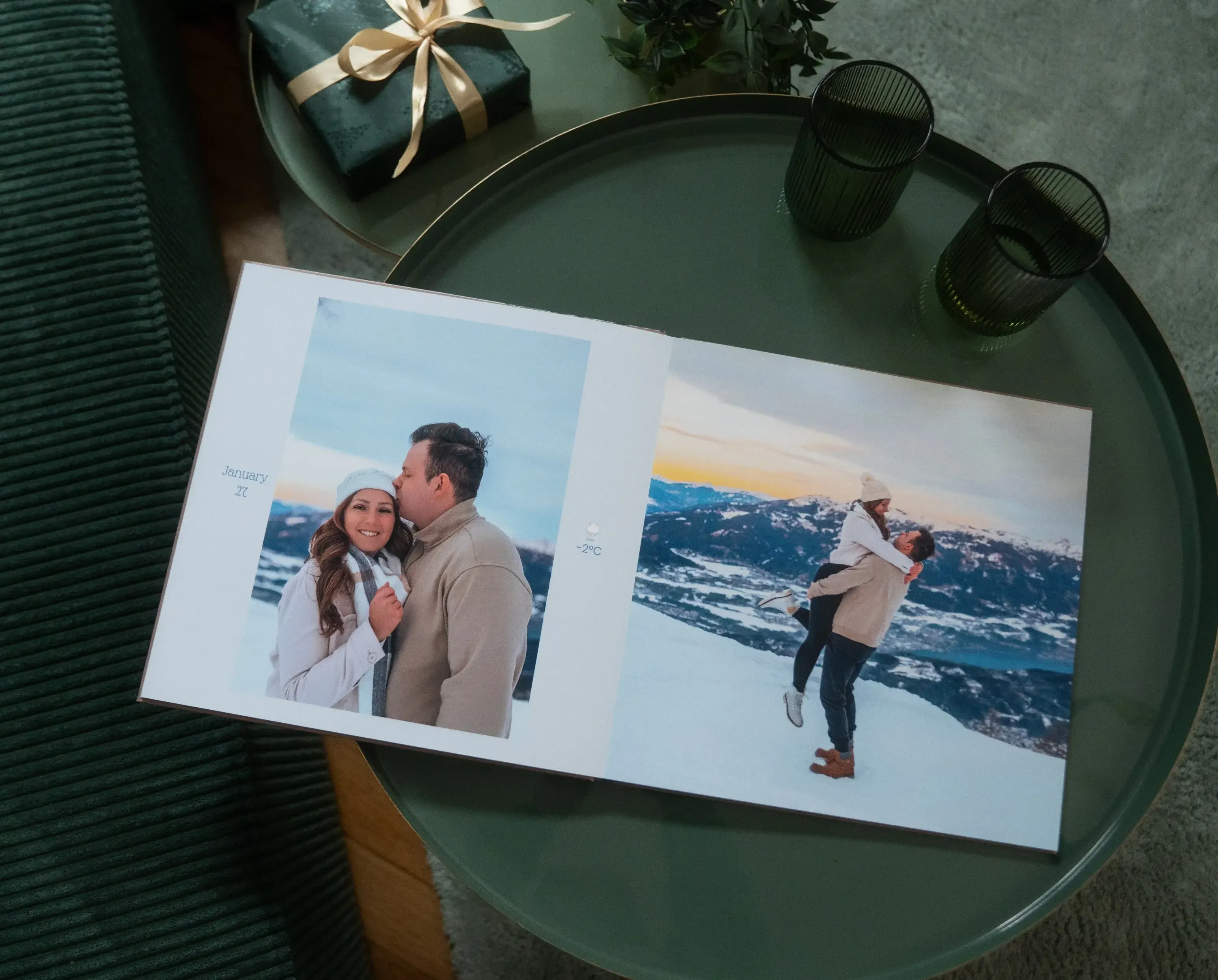 Open photo book on a green table, showing a couple in winter attire with snowy mountain backgrounds. Nearby are two green glasses and a wrapped gift.