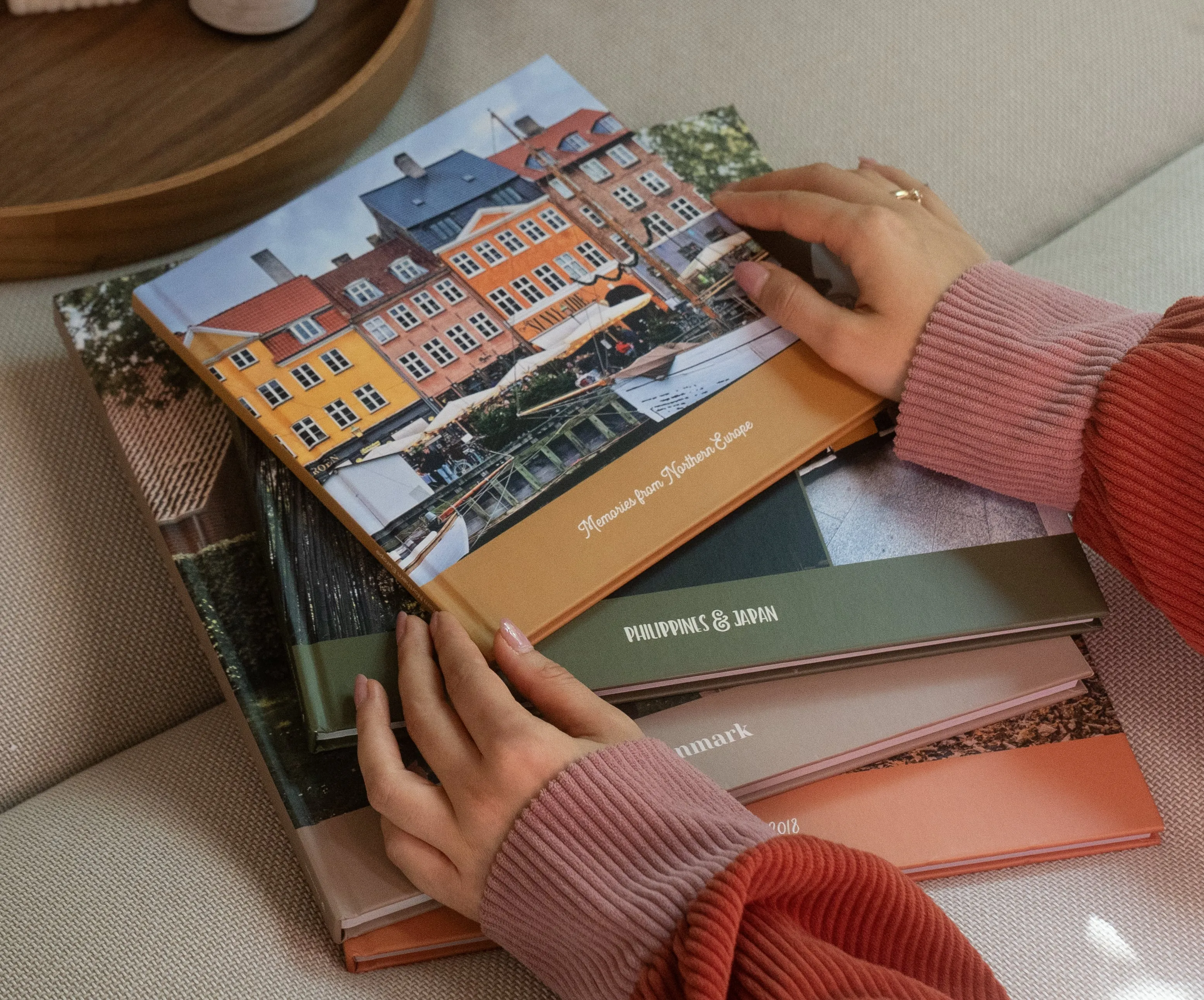 Hands holding a book with a cover showing colorful buildings by a canal, resting on a stack of books on a couch.