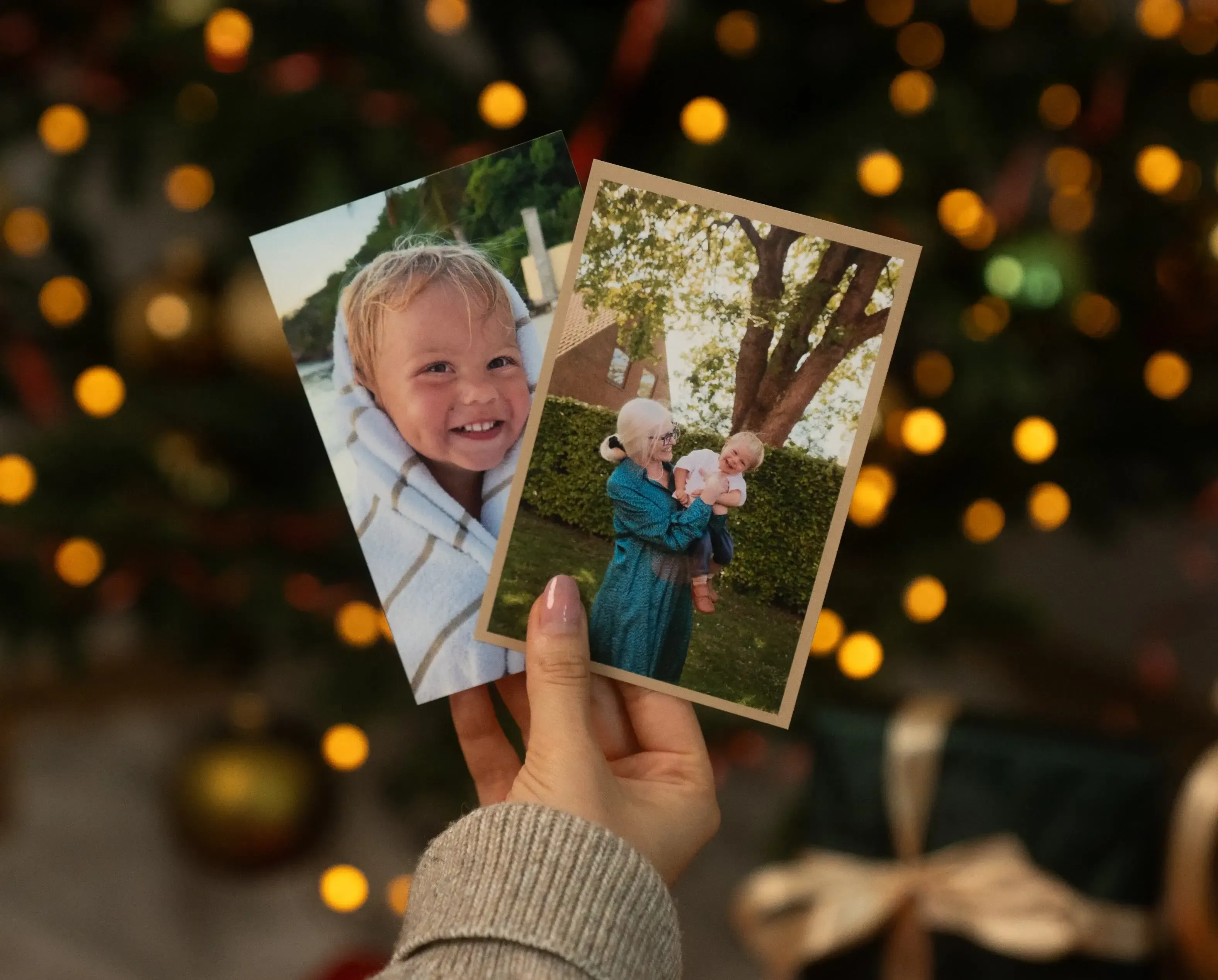 A hand holds two photos: a smiling child wrapped in a towel and an older woman lifting a child outdoors. Blurred Christmas lights in the background.