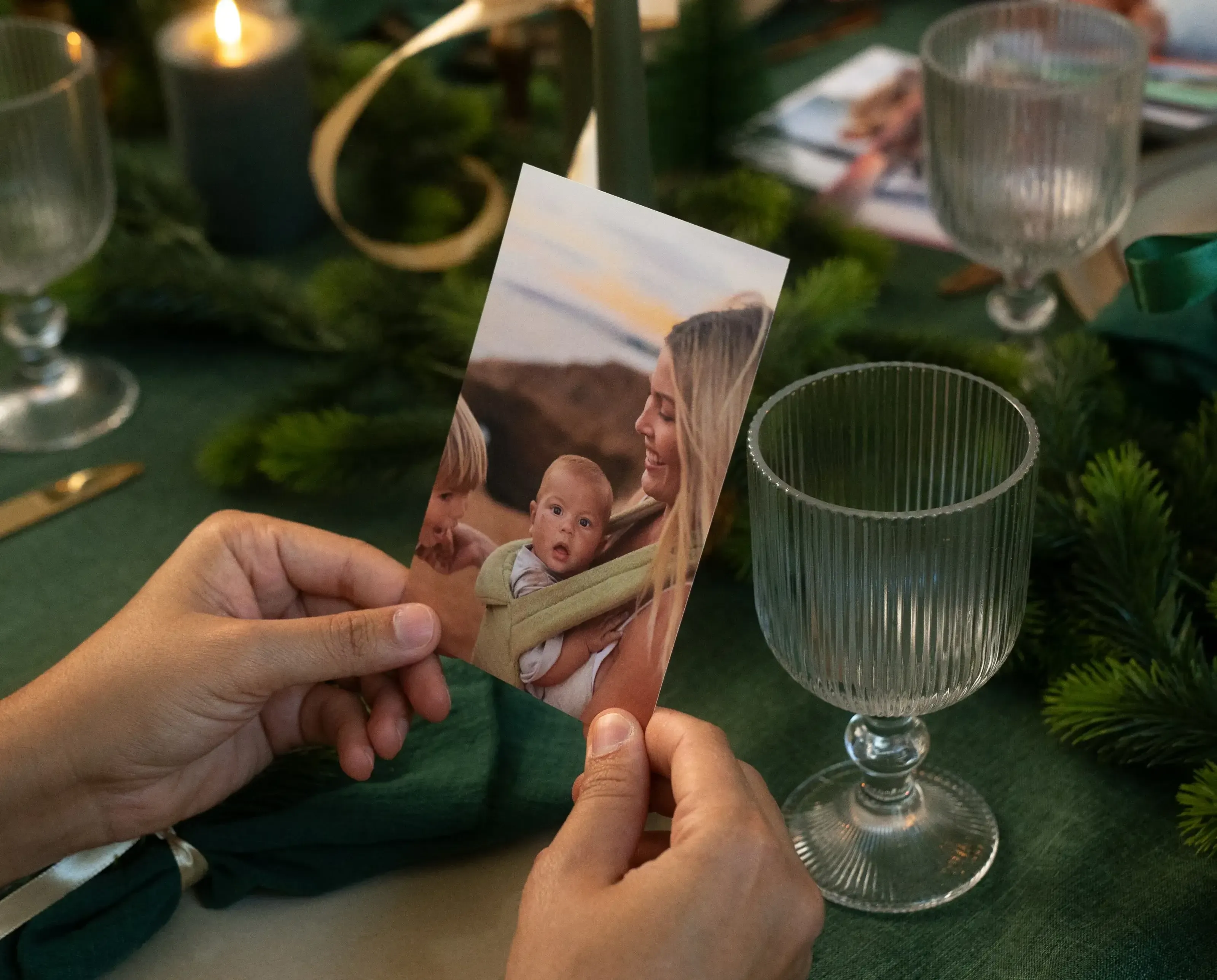 Hands holding a photo of a smiling woman with two children, set on a table with green decorations, candles, and glassware.