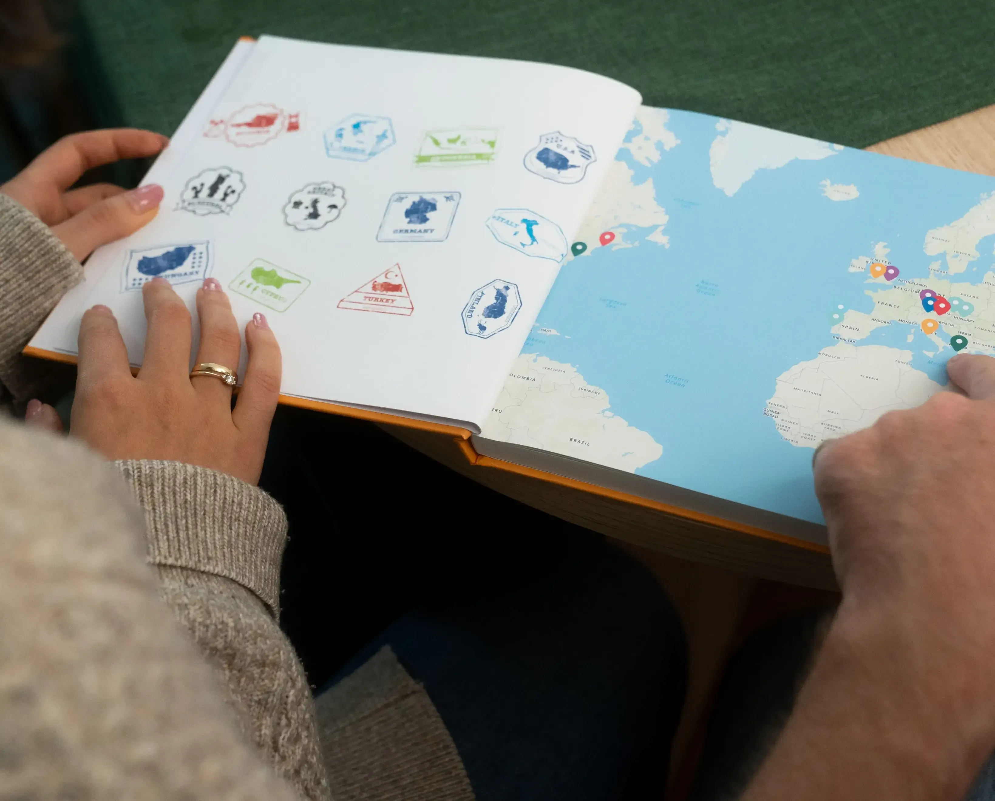 Two people look at a book featuring a world map and travel stamps. One person points to a location on the map.