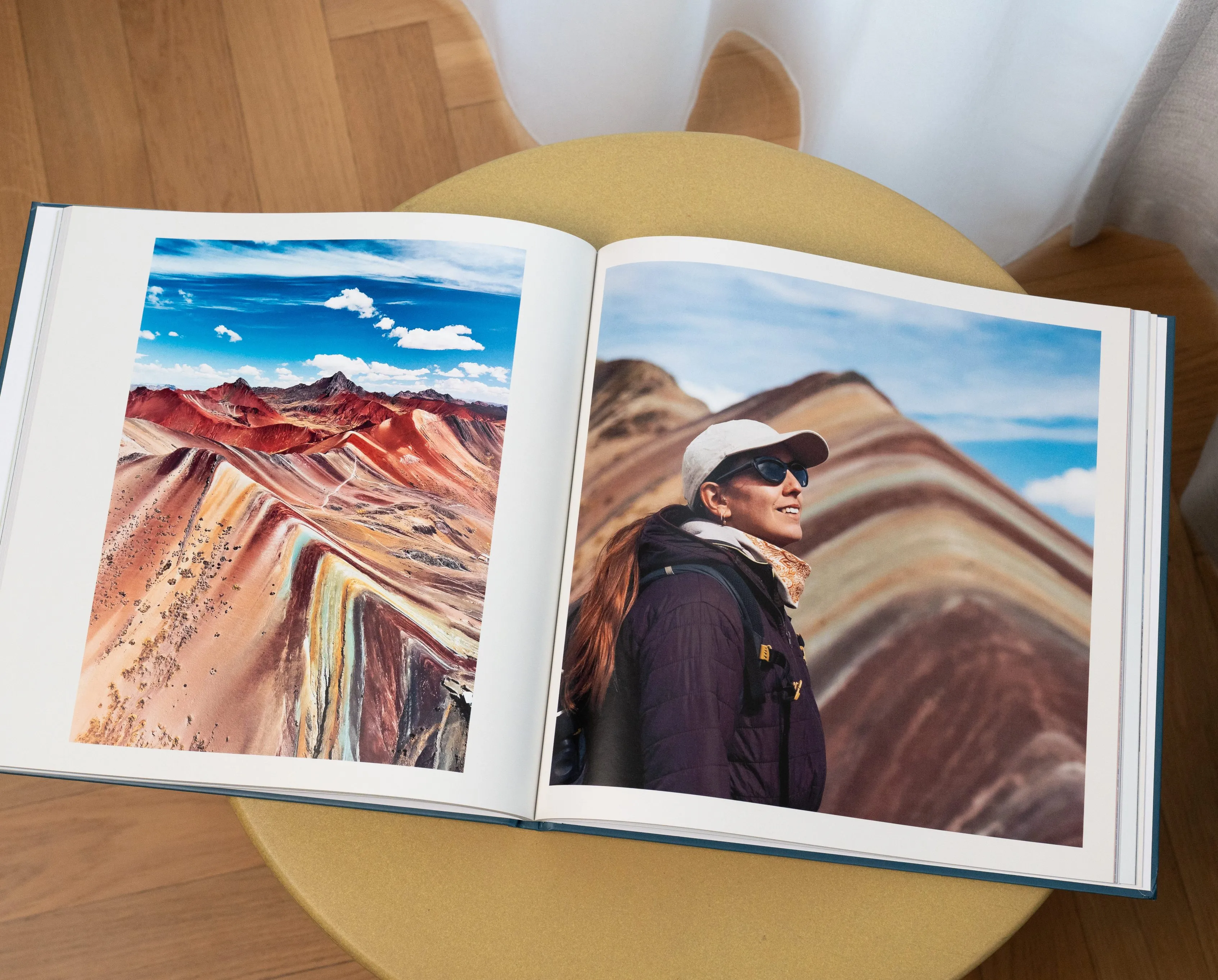 Ein aufgeschlagenes Reisefotobuch auf einem runden Tisch, auf dem Fotos einer Berglandschaft und einer Person mit Mütze und Sonnenbrille zu sehen sind.