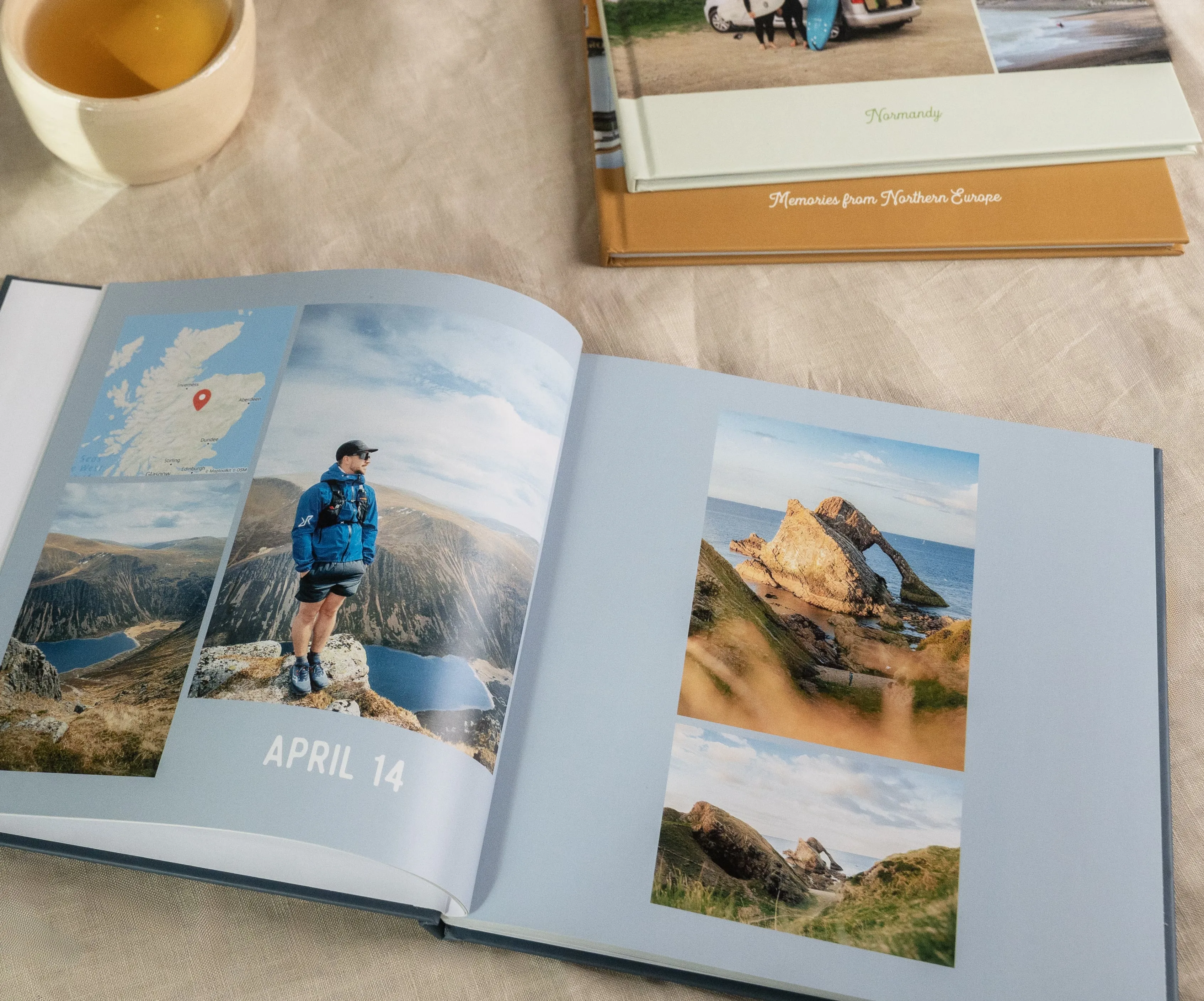 Open photo book on a table, showing travel photos of landscapes and a person hiking. A cup and more books are nearby.