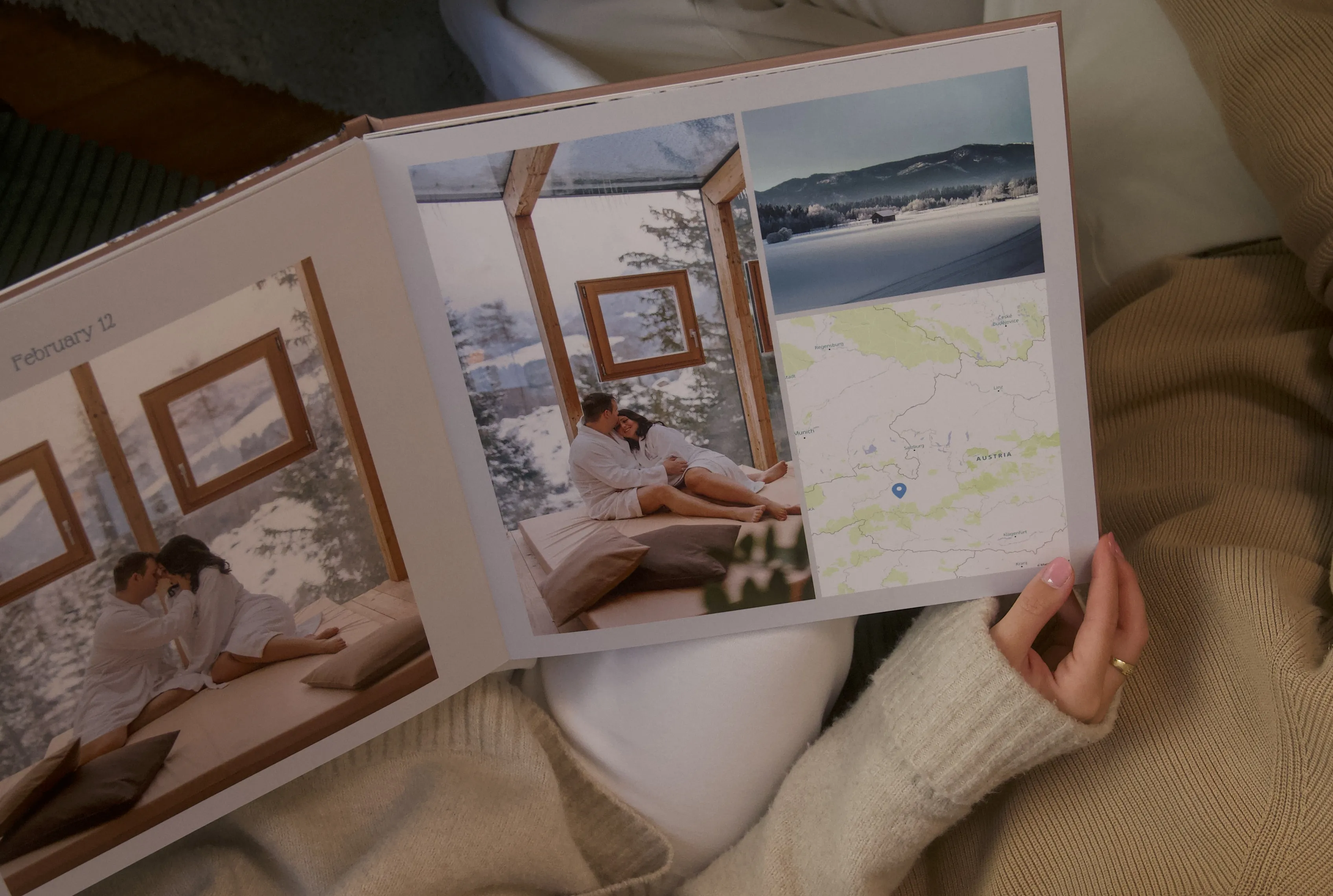 Person holding an open photo album with winter cabin photos and a map, lying on a cozy blanket.