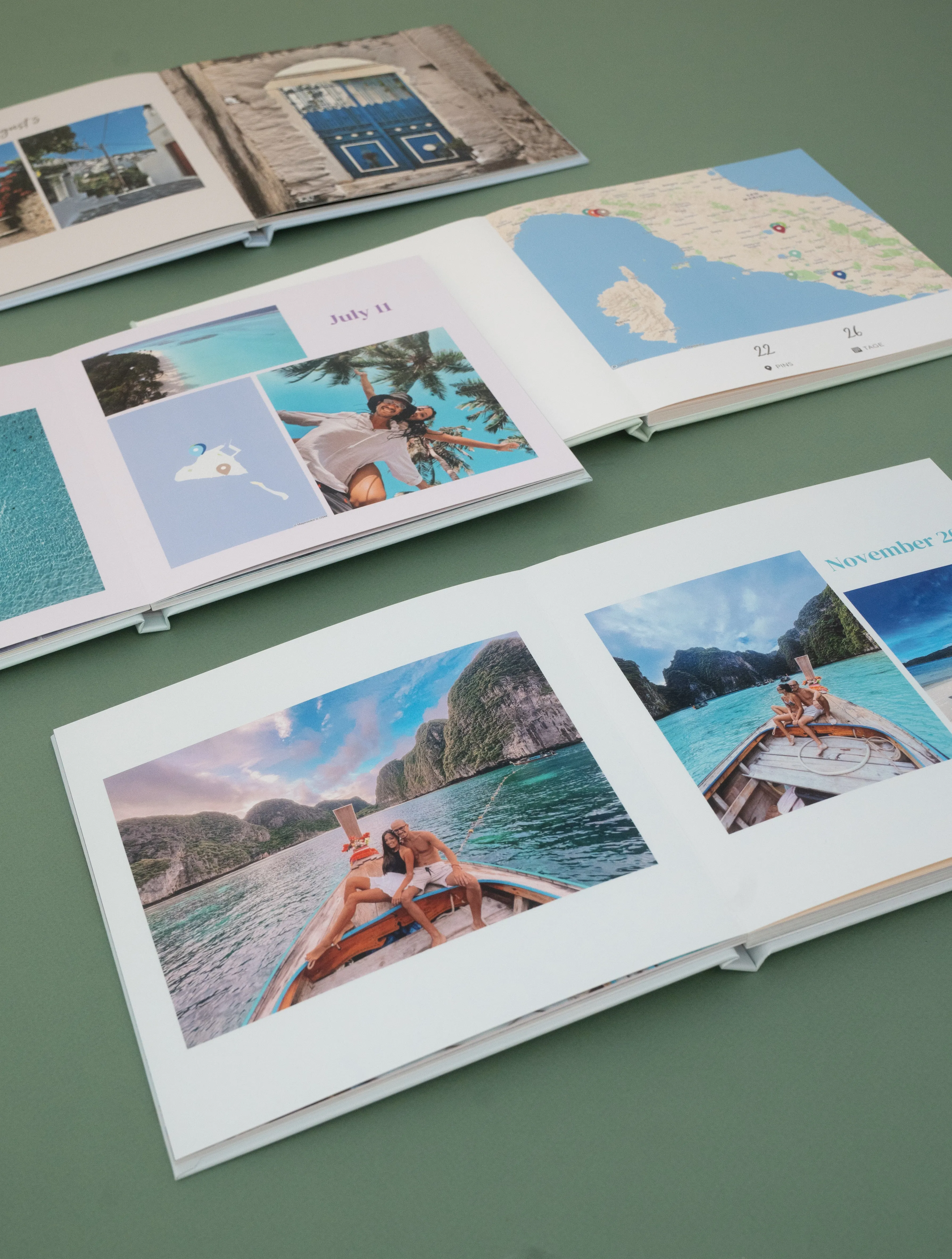 Open travel photo albums on a green surface, featuring travel photos, maps, and dates like July 11 and November 7.
