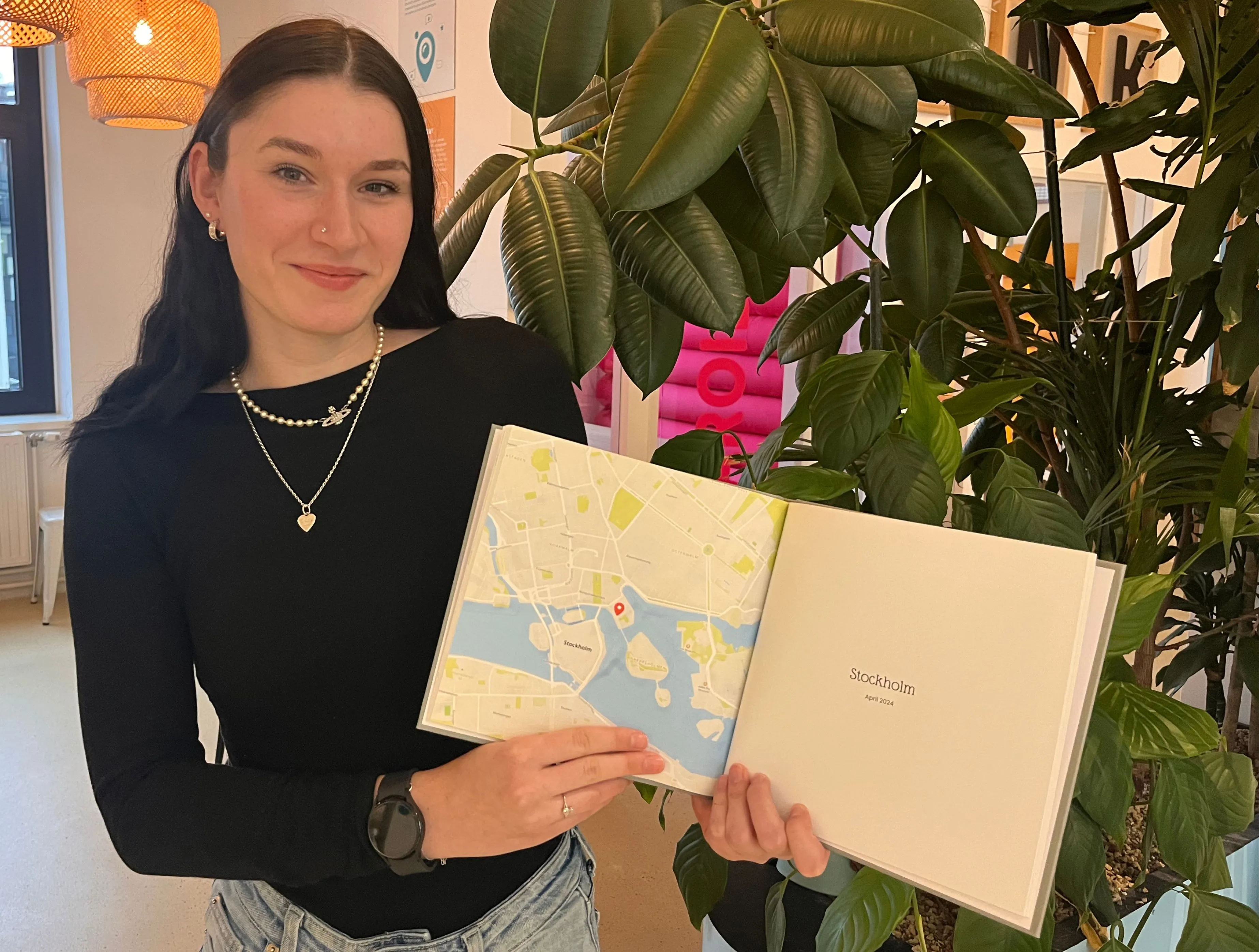 Woman in black shirt holds open a photo book displaying a map, standing next to a large indoor plant.