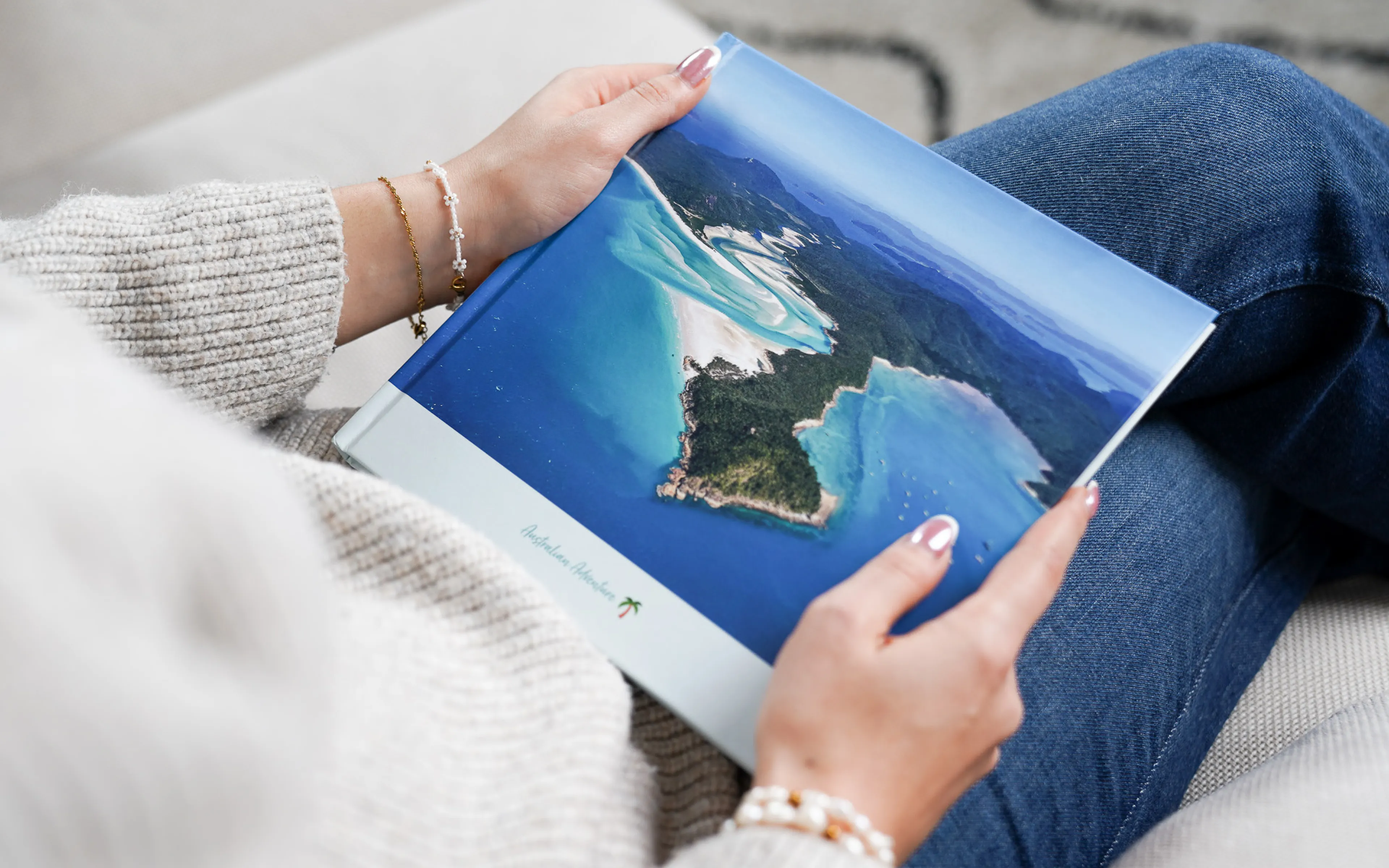Person holding a book with an aerial photo of a tropical island and turquoise waters, wearing a gray sweater and jeans.