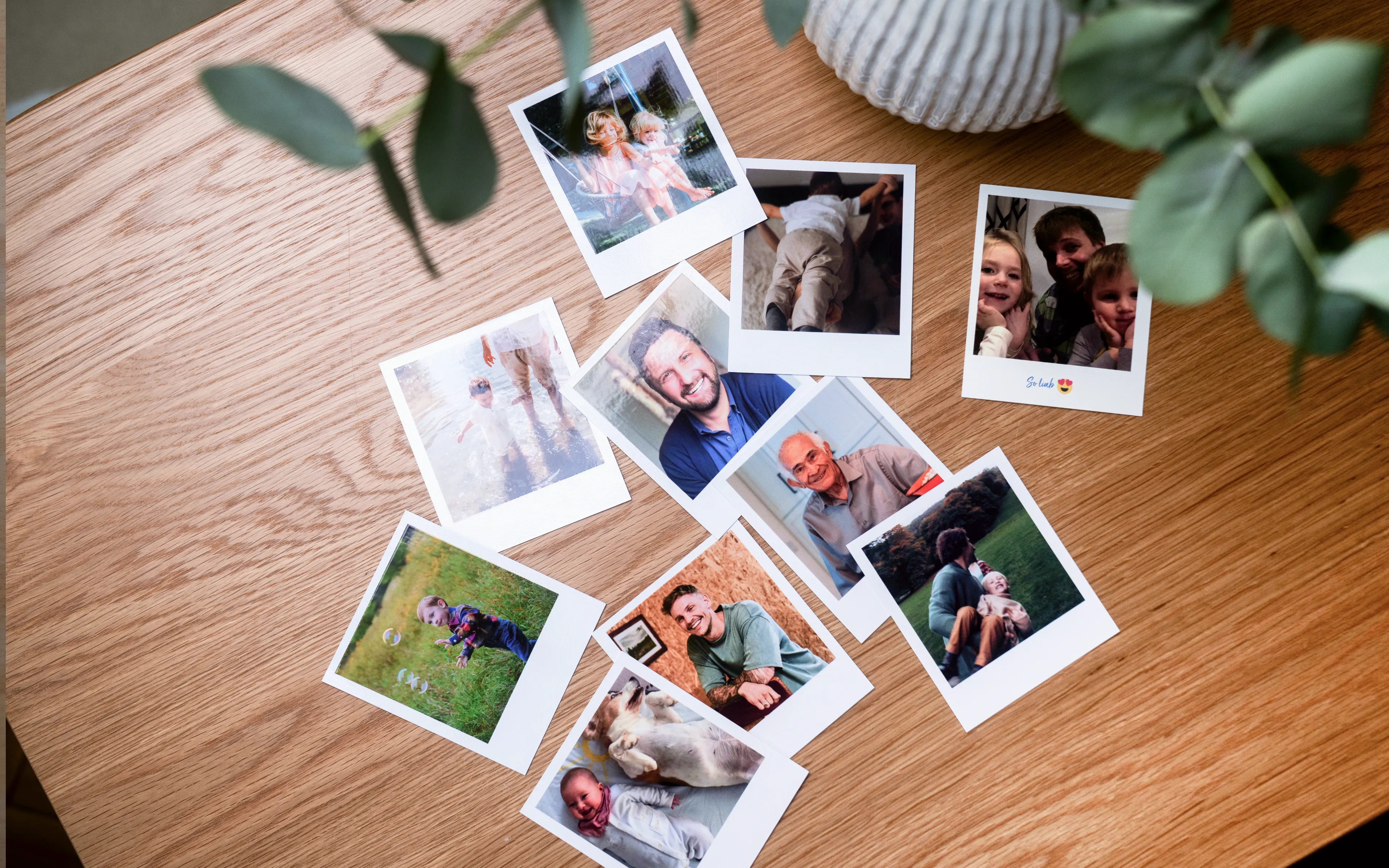 A collection of Polaroid photos scattered on a wooden table, featuring various people and moments. A plant is partially visible in the corner.