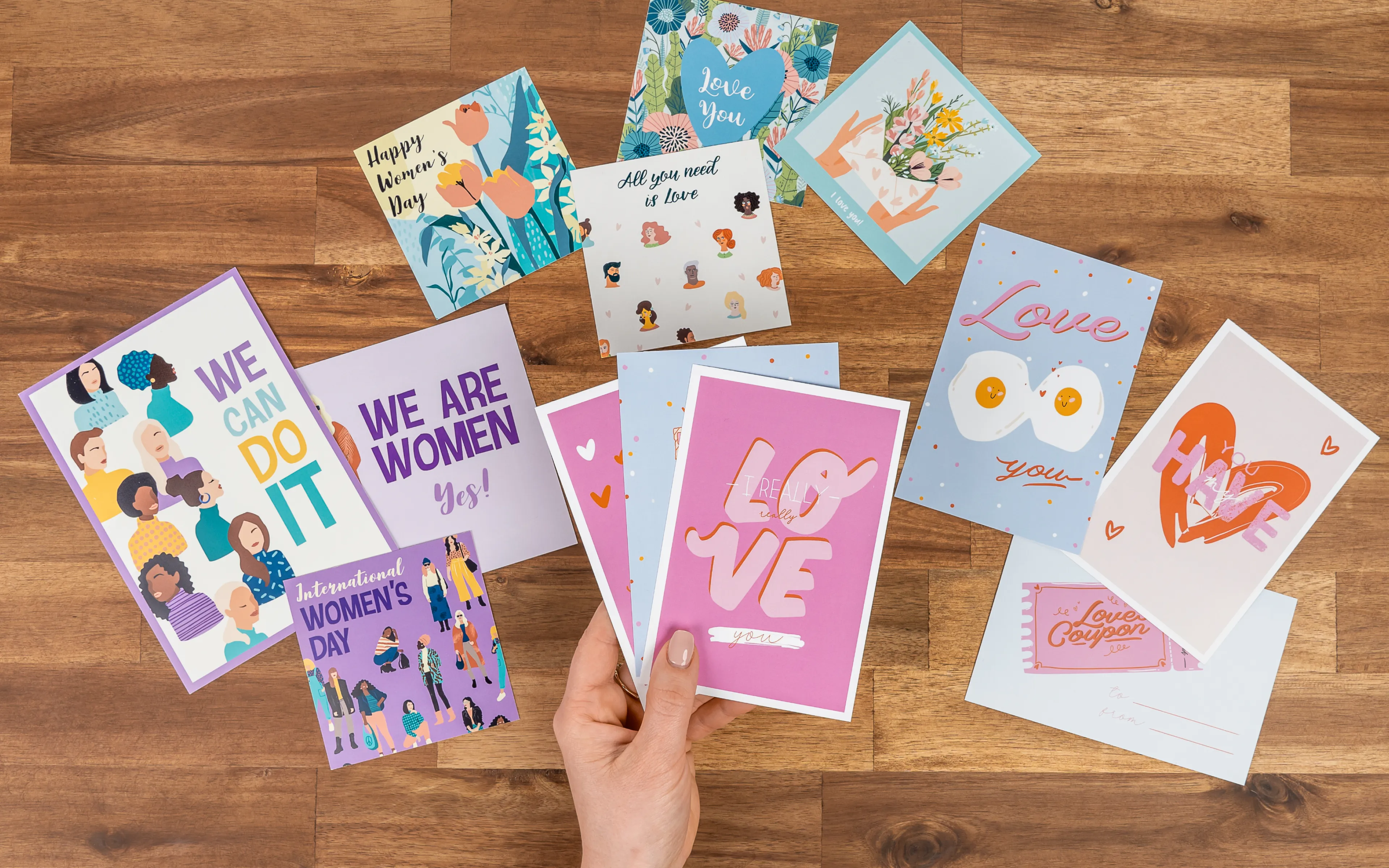 A hand holds a pink "LOVE" card surrounded by various colorful greeting cards on a wooden surface.