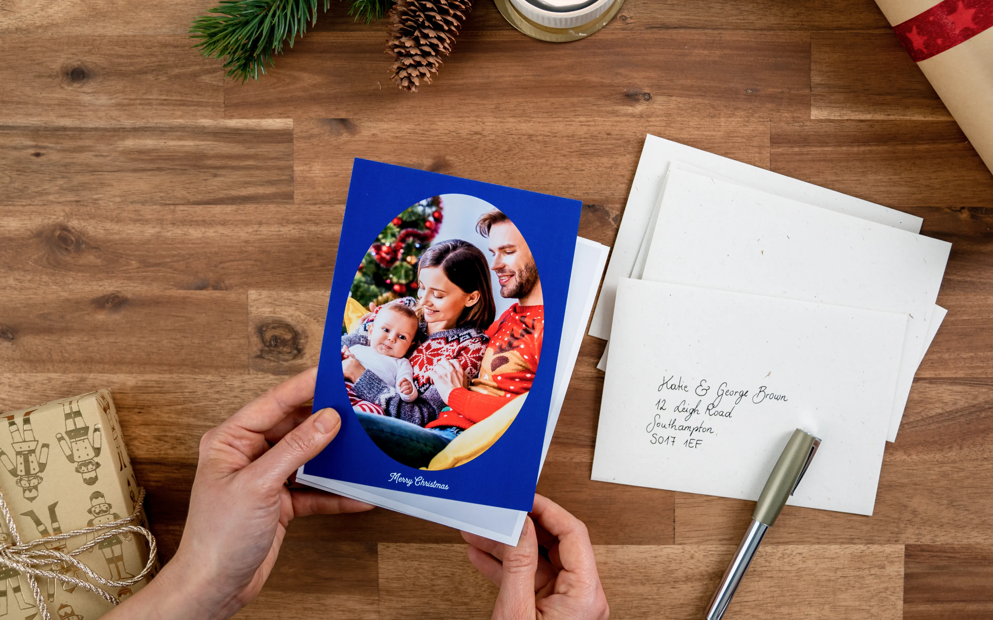 Hands holding a Christmas card with a family photo, surrounded by envelopes, a pen, and a wrapped gift on a wooden table.