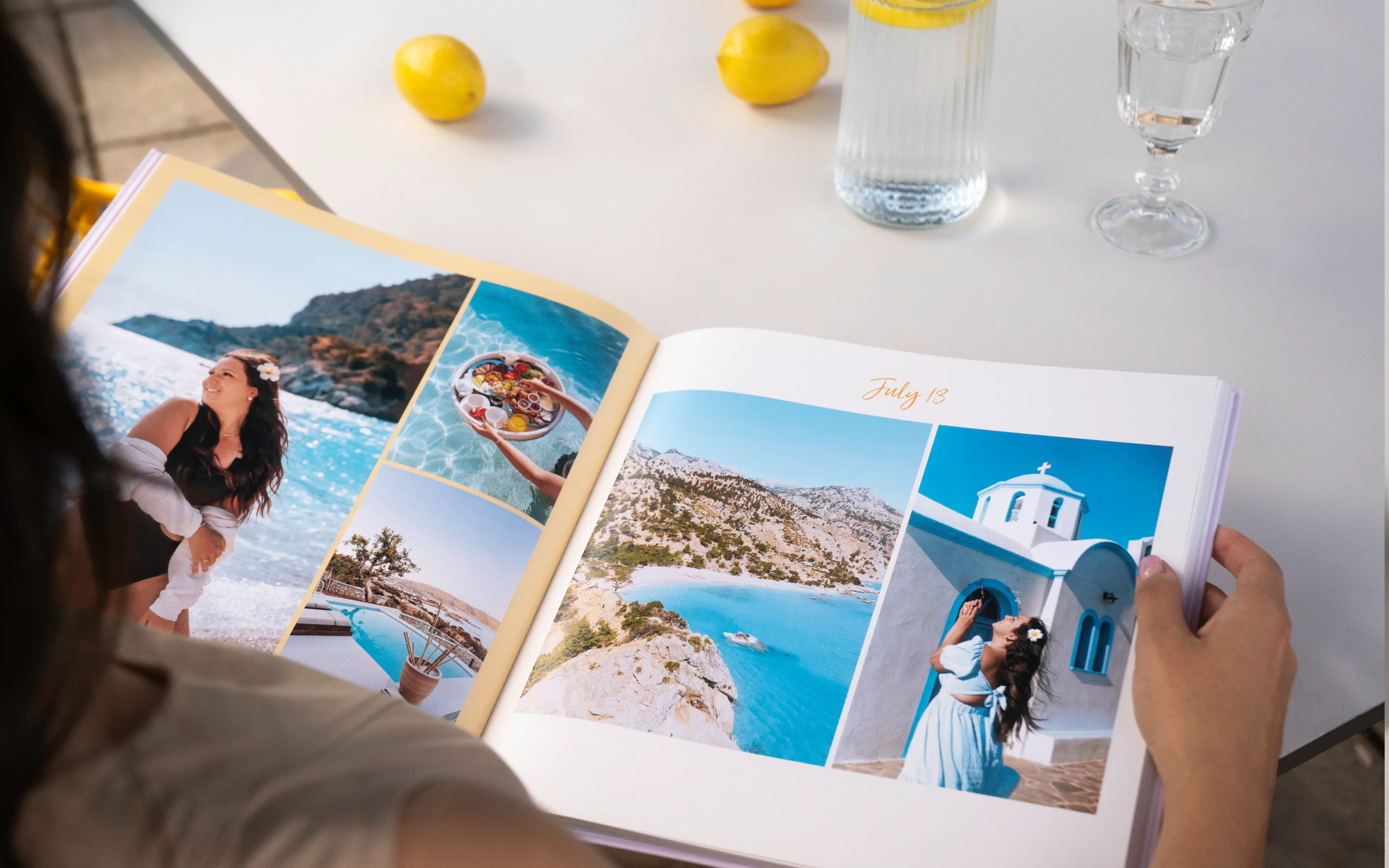Person viewing a travel photo book with images of a coastal vacation, featuring beaches and a woman in a blue dress. Lemons and drinks on the table.