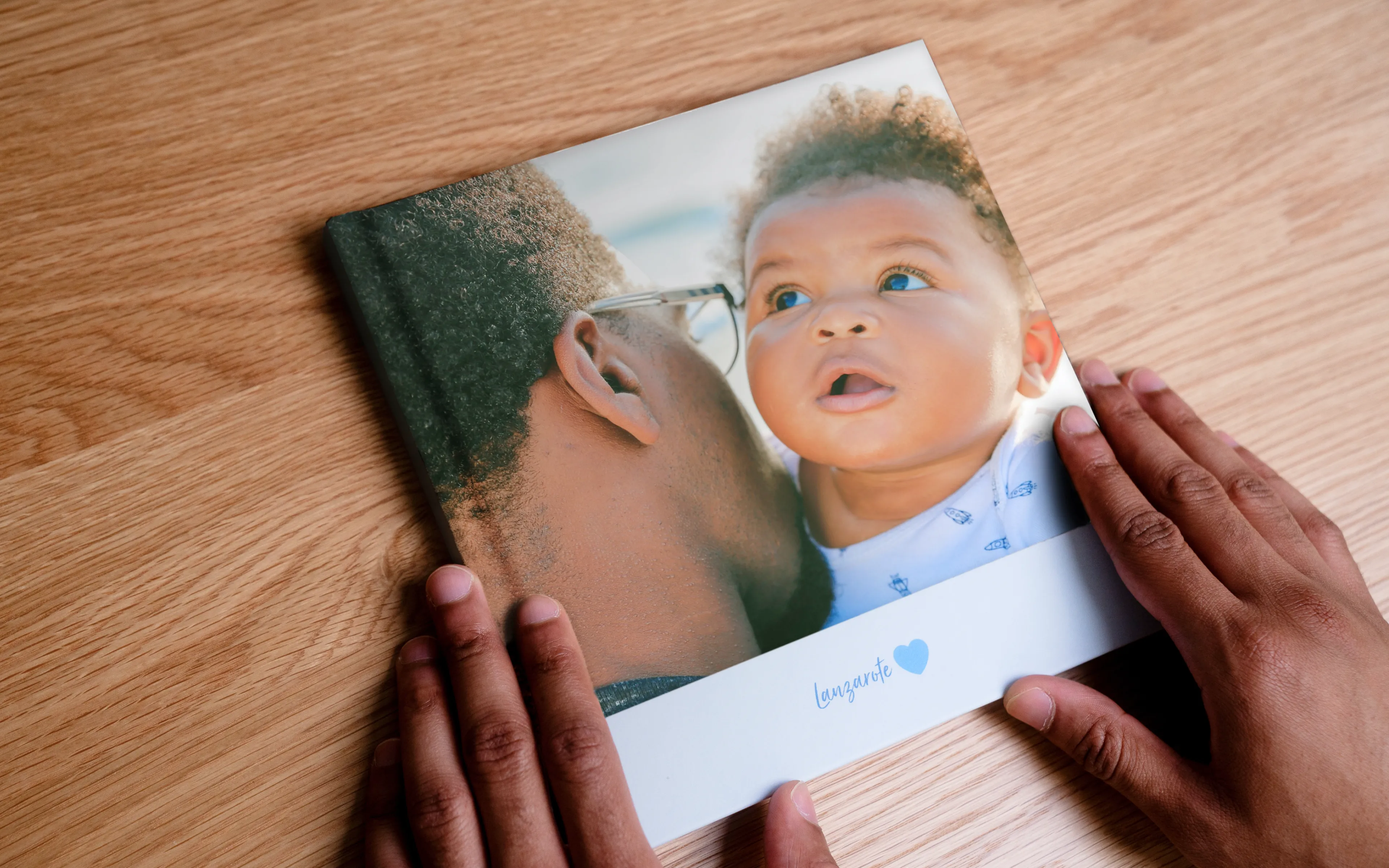Hände halten ein Fotobuch mit einem Cover, auf dem ein Mann ein Baby mit lockigem Haar auf die Wange küsst, auf einer Holzoberfläche.
