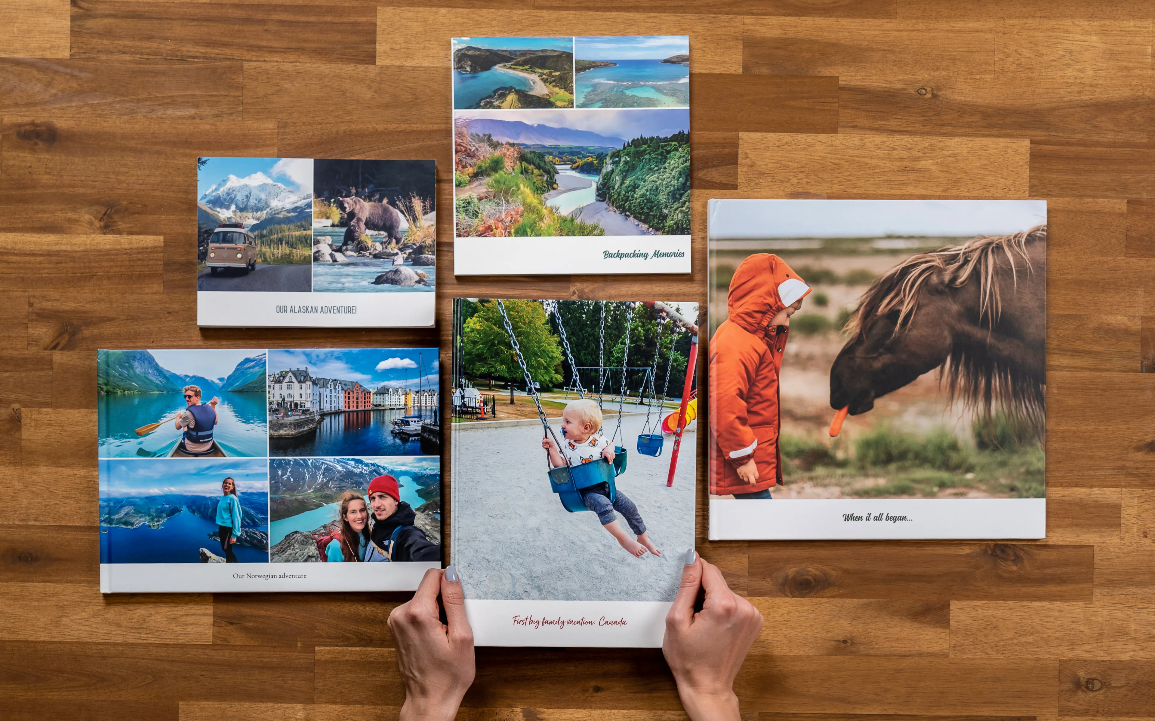 Hands arranging five travel photo books on a wooden table, featuring scenic landscapes, a child on a swing, and a horse with a carrot.