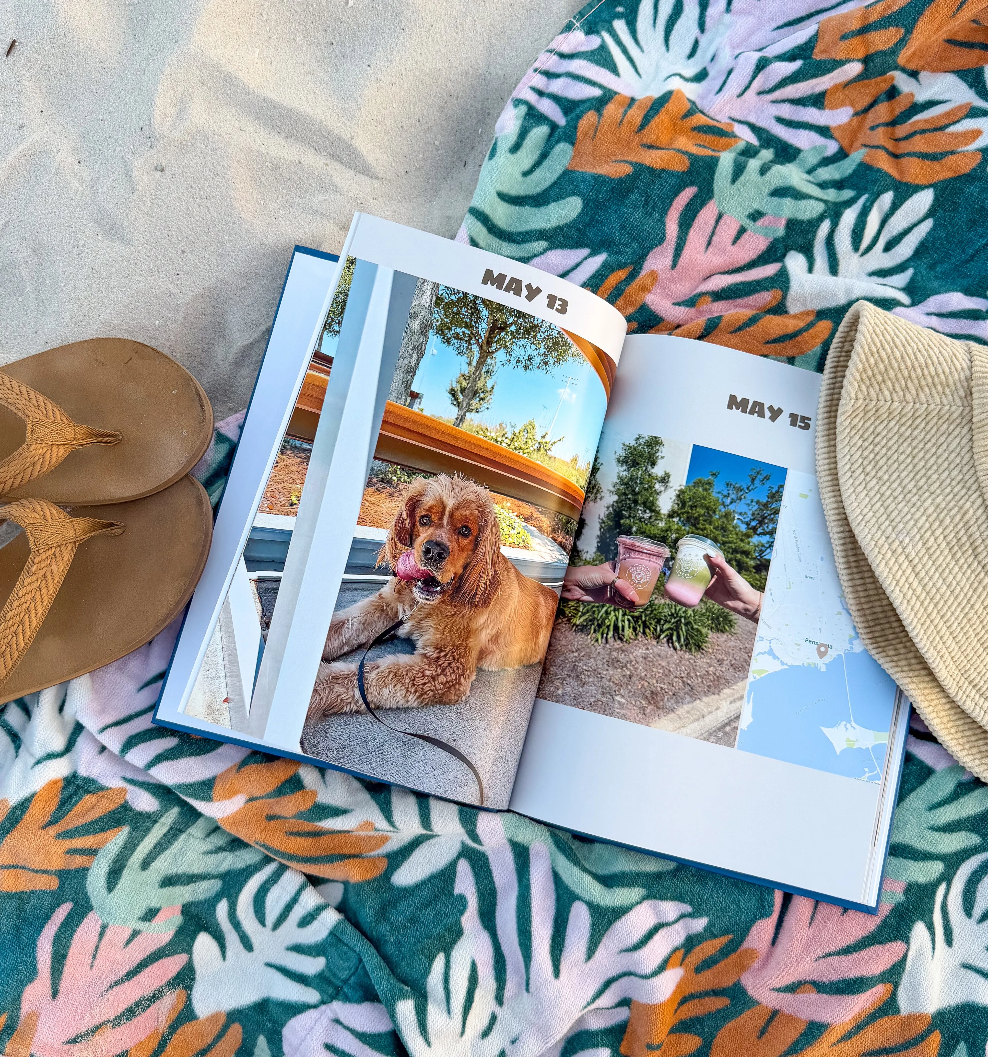 Open book with photos of a dog and drinks on a tropical-patterned towel, next to sandals and a straw hat on sand.