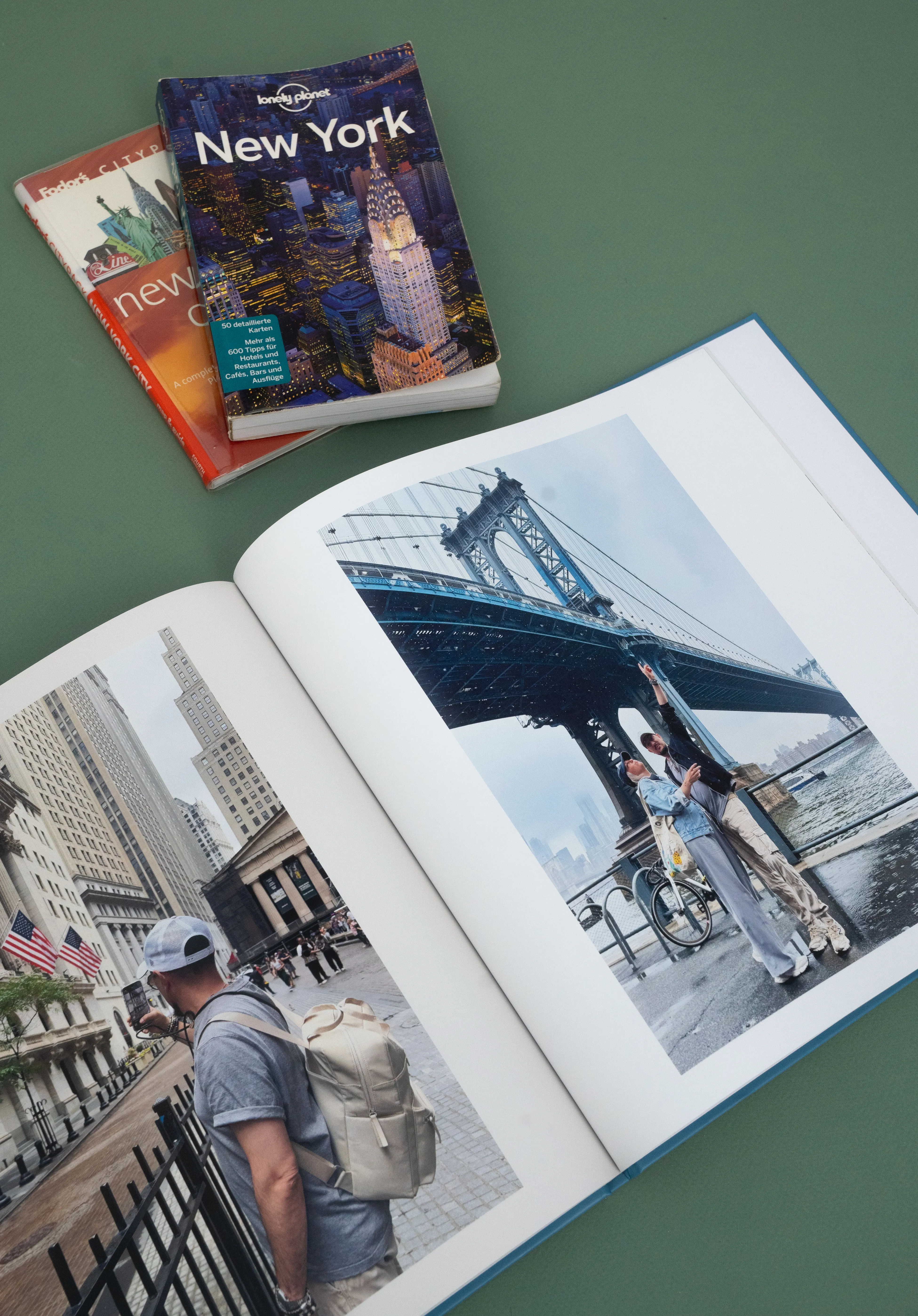 Offenes Softcover-Fotobuch mit Bildern von Sehenswürdigkeiten in New York City, darunter eine Brücke und eine Straßenszene. Zwei Reiseführer liegen daneben auf einer grünen Fläche.
