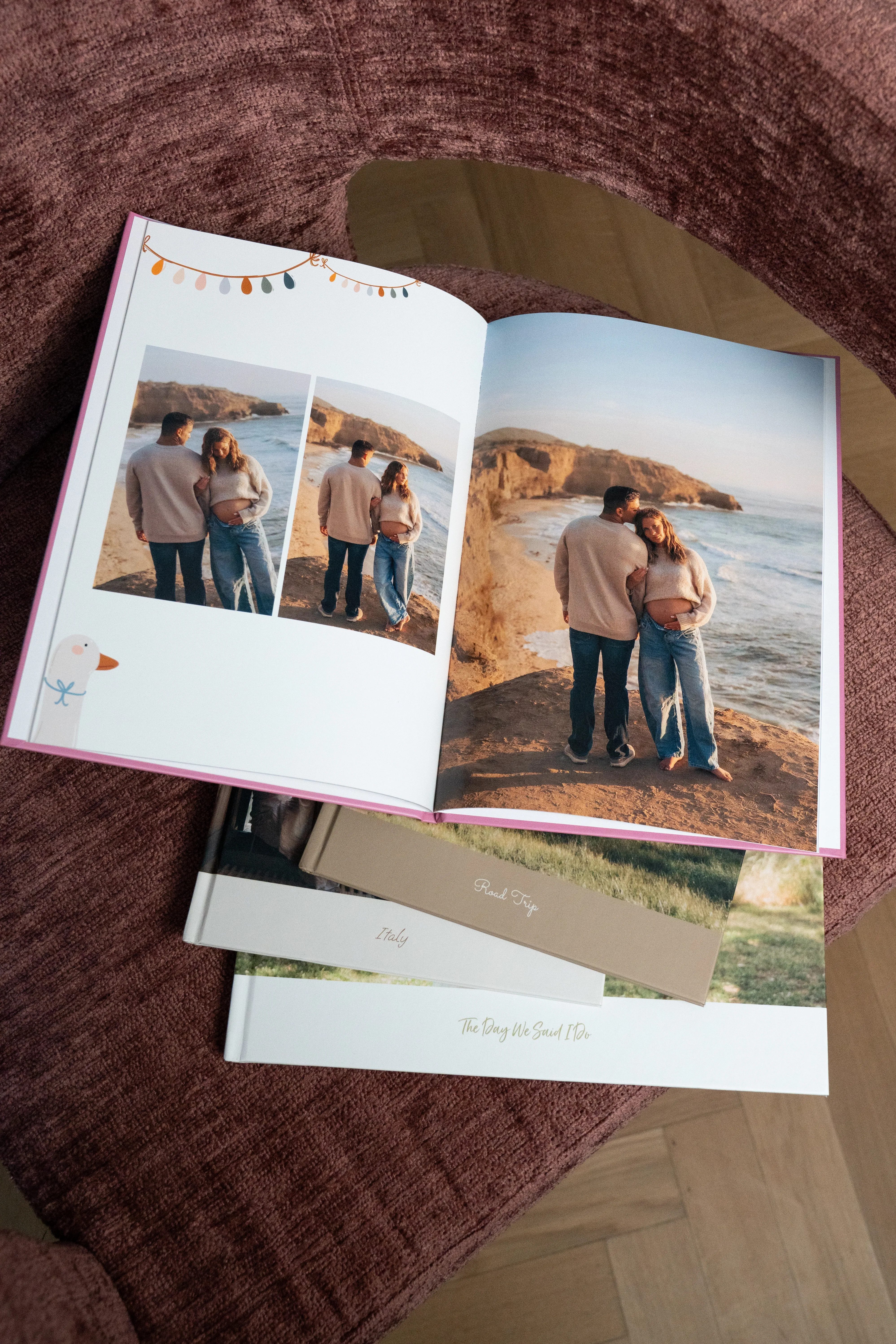 Ein aufgeschlagenes Fotobuch, das ein Paar am Meer zeigt, darunter liegen weitere Bücher auf einer strukturierten braunen Oberfläche gestapelt.