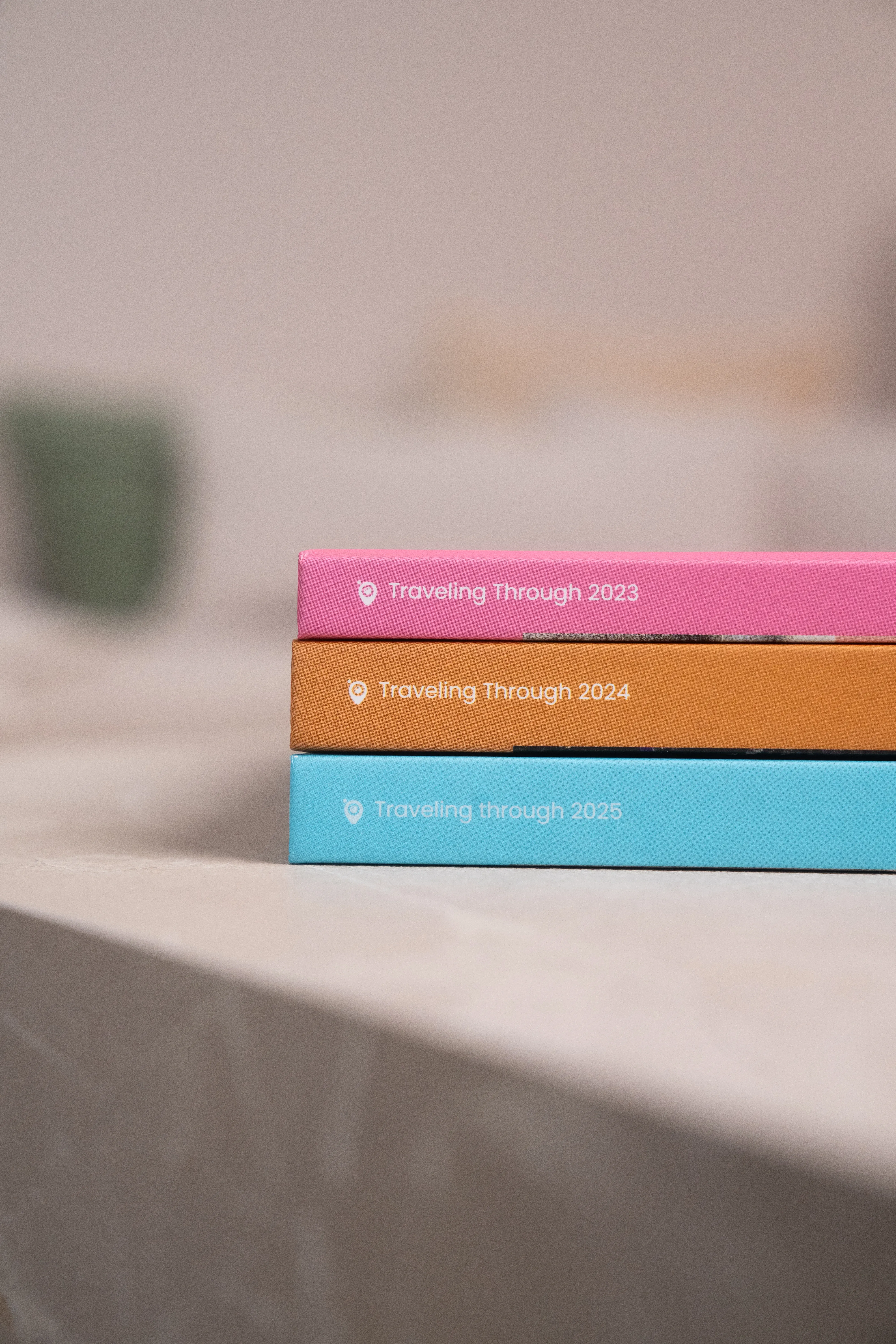 Trois carnets de voyage empilés — rose « Voyager à travers 2023 », orange « Voyager à travers 2024 », bleu « Voyager à travers 2025 » — sur une surface neutre.