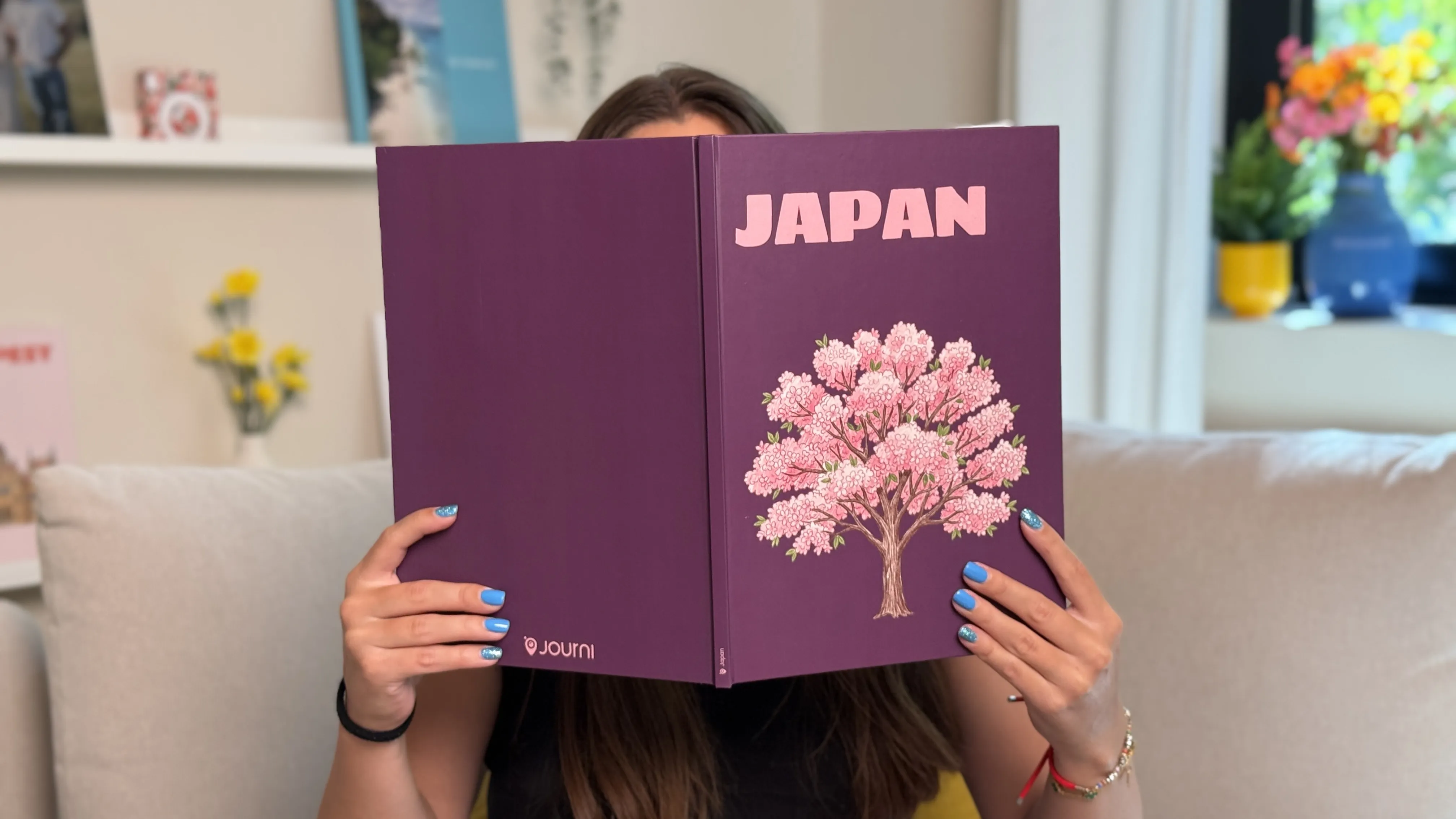 A person with painted blue nails holds a custom photo book titled "JAPAN" with a cherry blossom image, seated indoors near a window and flowers.