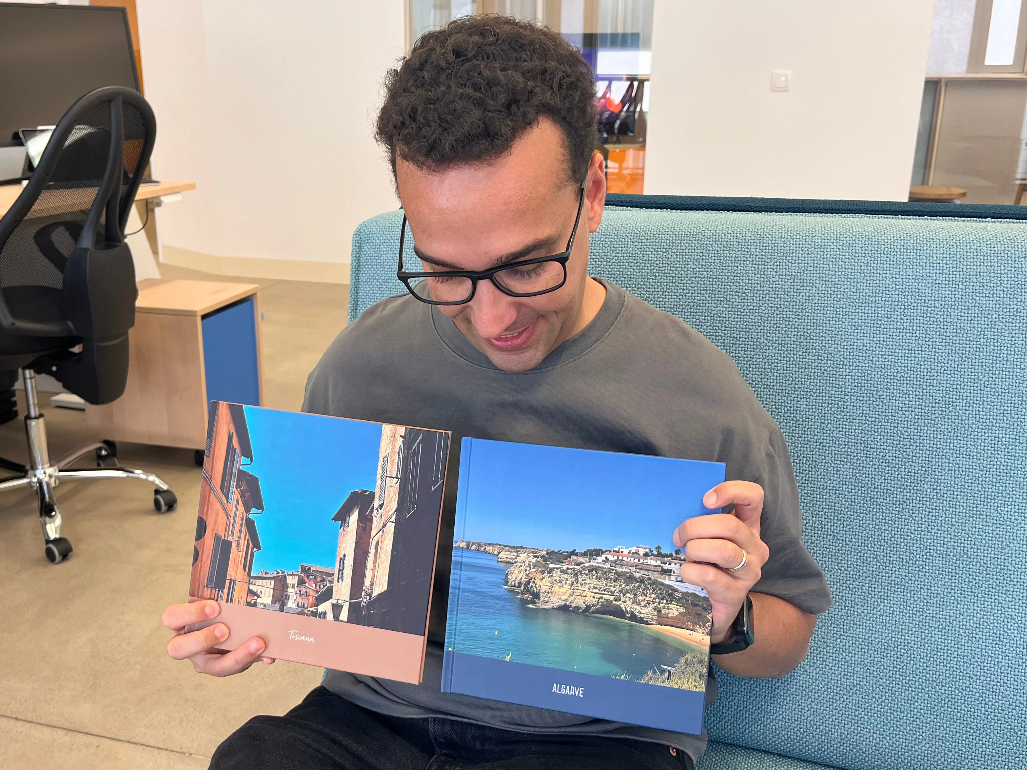 A person sitting on a couch holds two open photo books, showing images of colorful buildings and a coastal landscape.