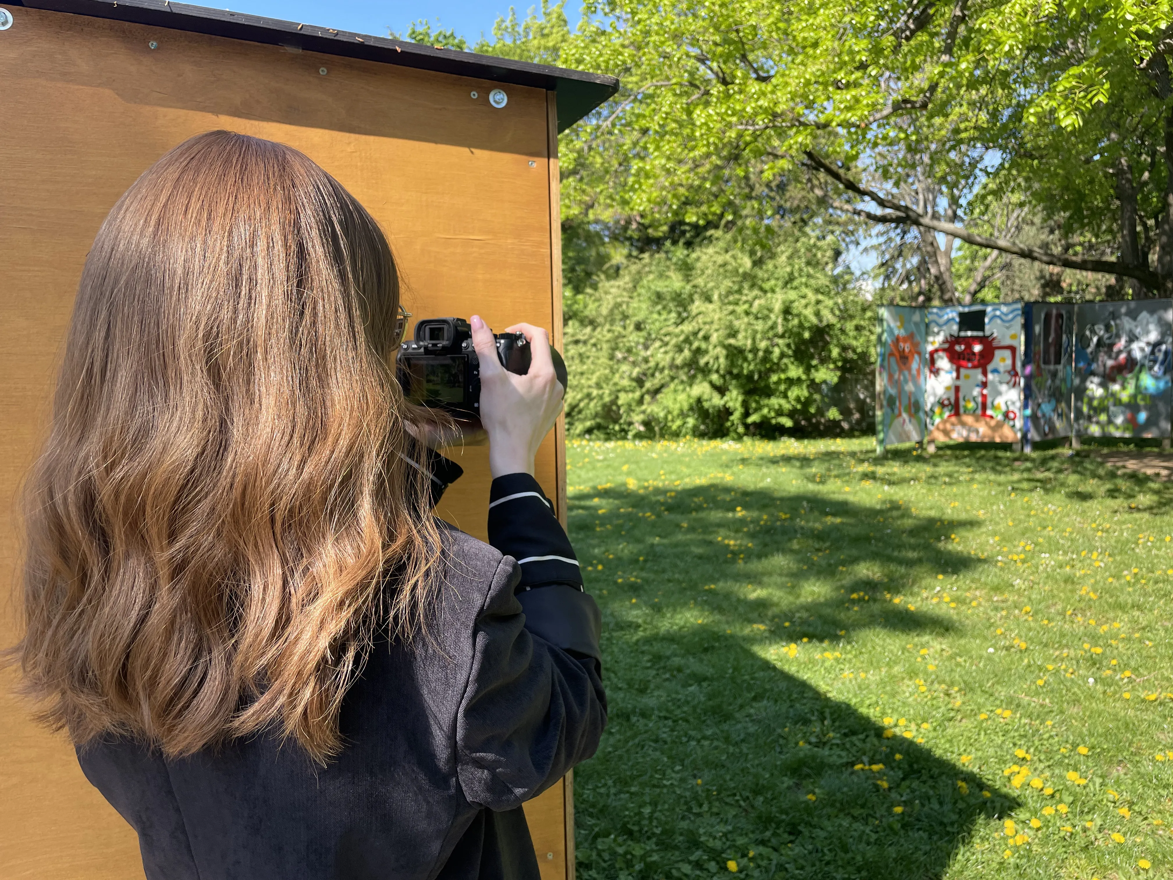 Person with long hair photographing a colorful graffiti wall in a sunny park, standing next to a wooden structure.