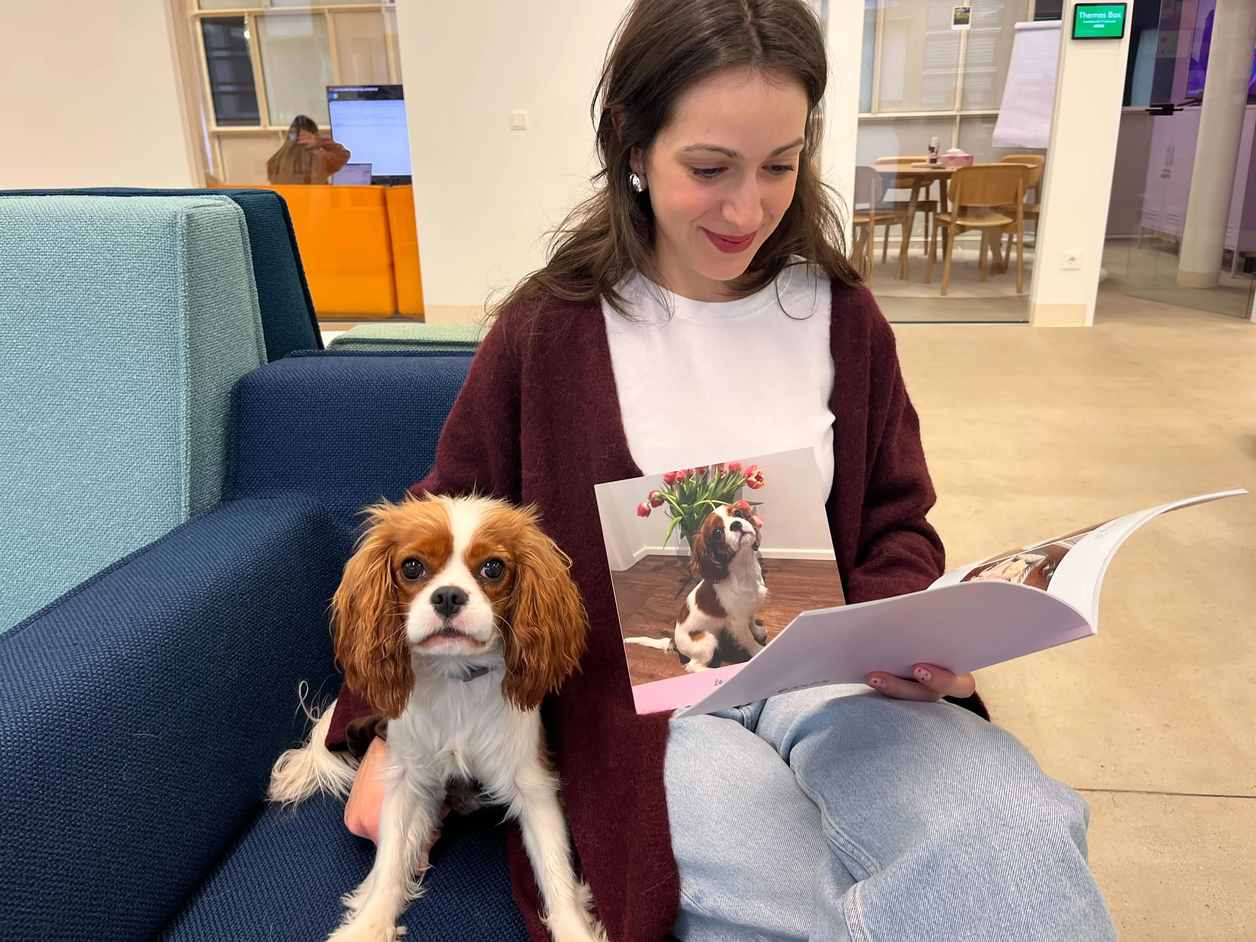 Woman sitting on a blue couch, reading a pet photo book with a Cavalier King Charles Spaniel beside her.
