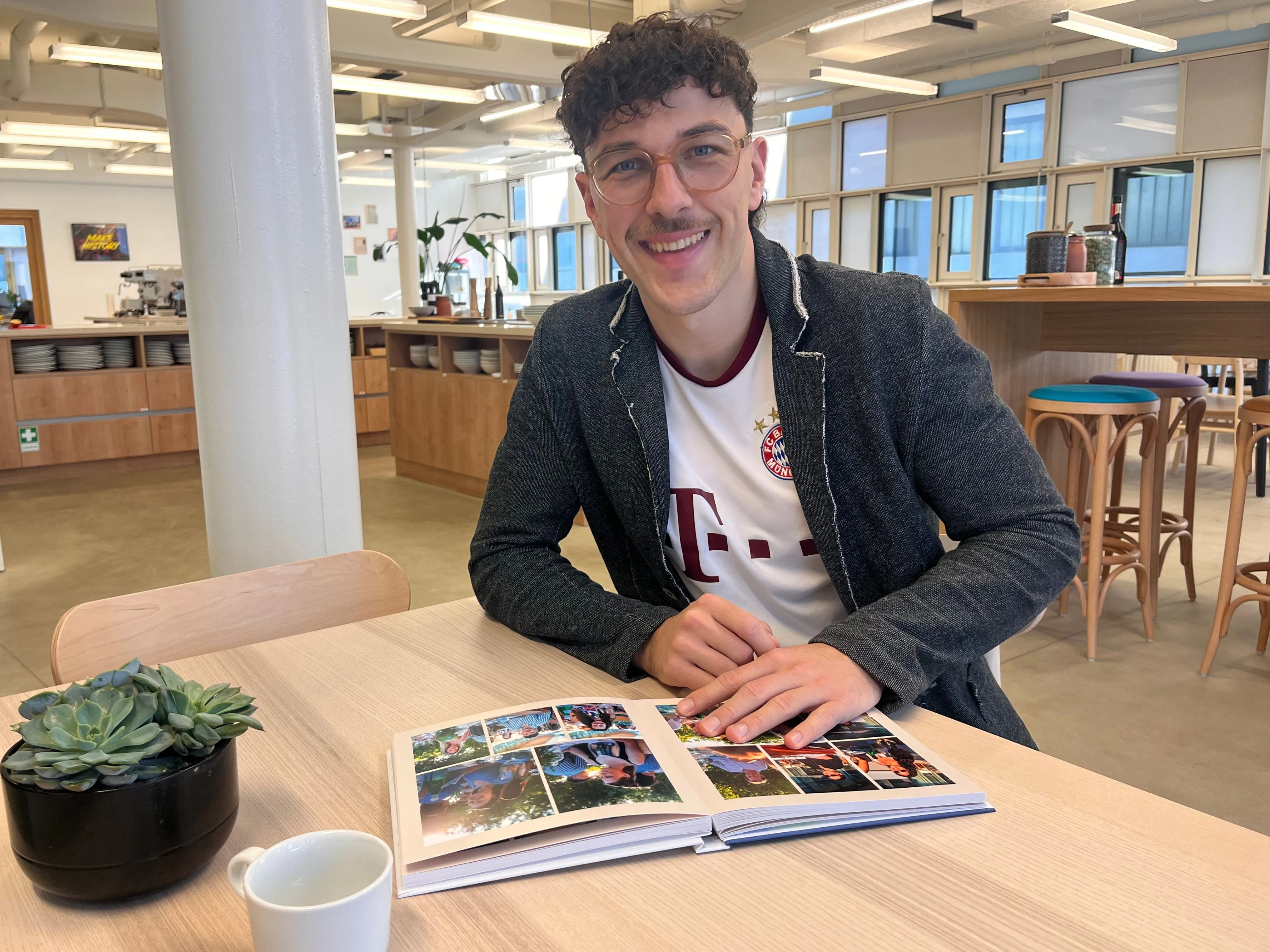 Smiling person with glasses and curly hair sits at a table, looking at a photo album. A succulent and a cup are nearby.