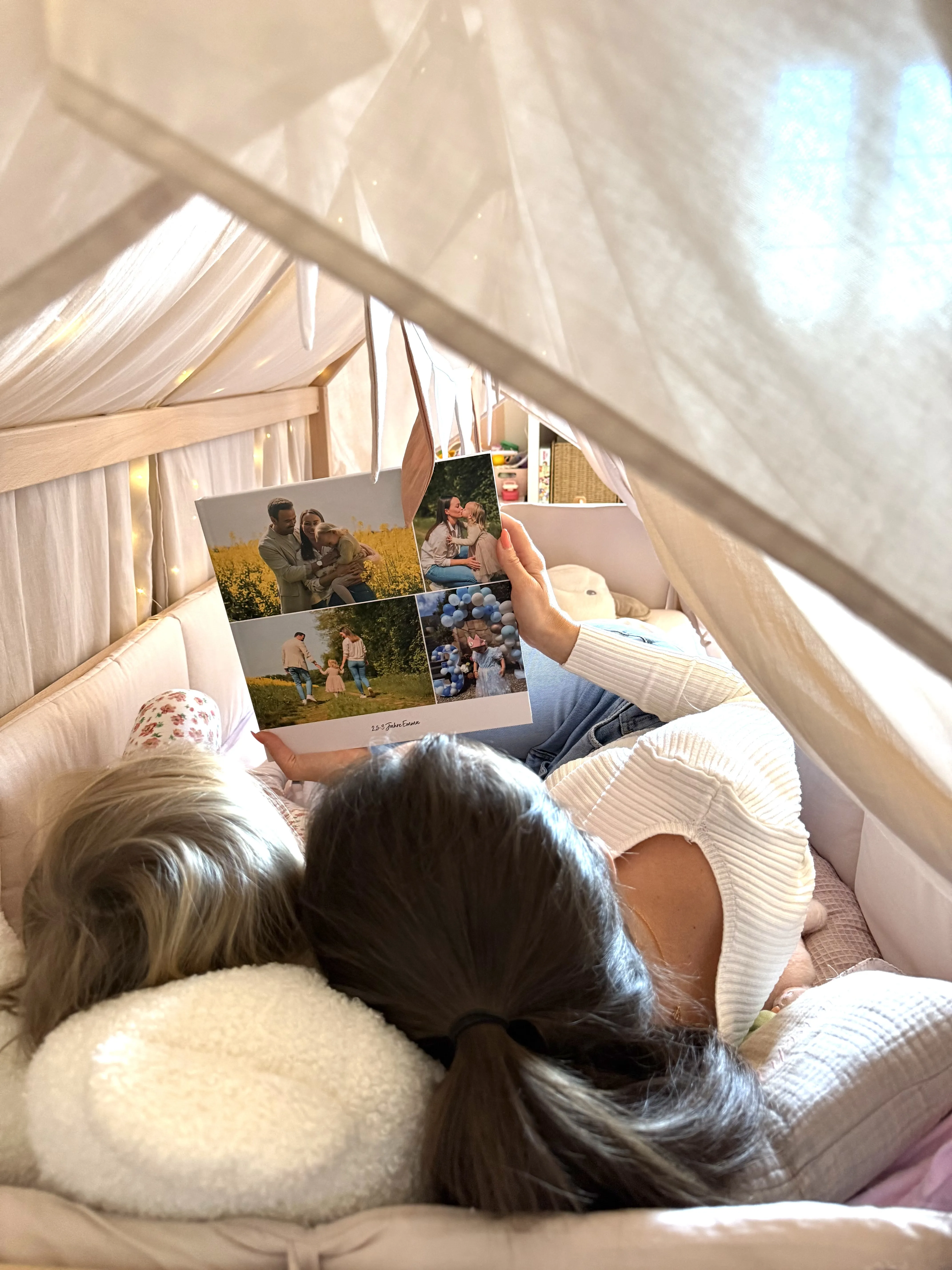 Anne und Tochter Emma schauen sich ein Fotobuch mit Fotos von Familienurlauben und Mama-Momenten in einem Kinderbett an.