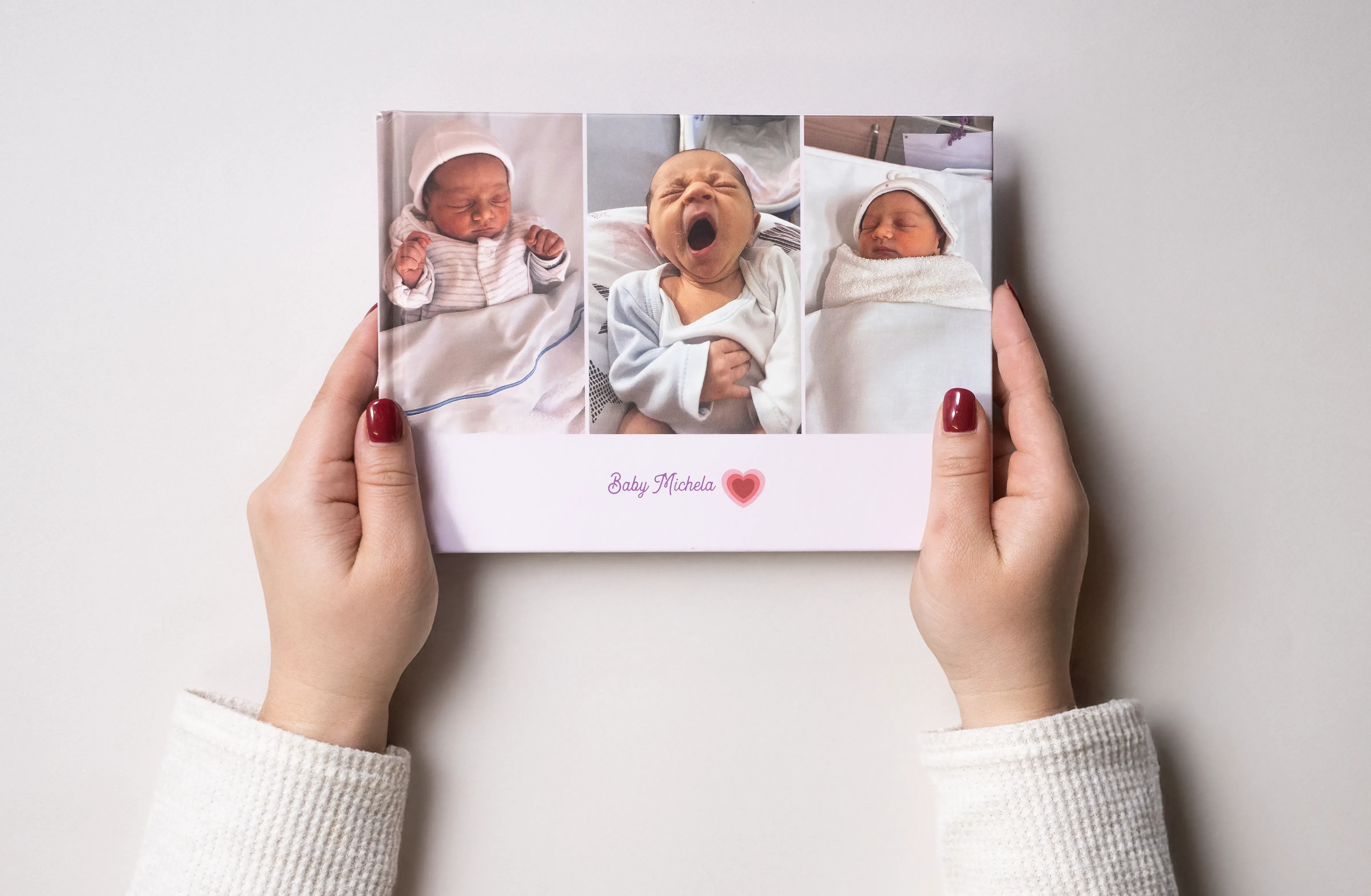 Hands holding a baby photo album with three pictures of a newborn baby in different poses, including yawning, on a light background.