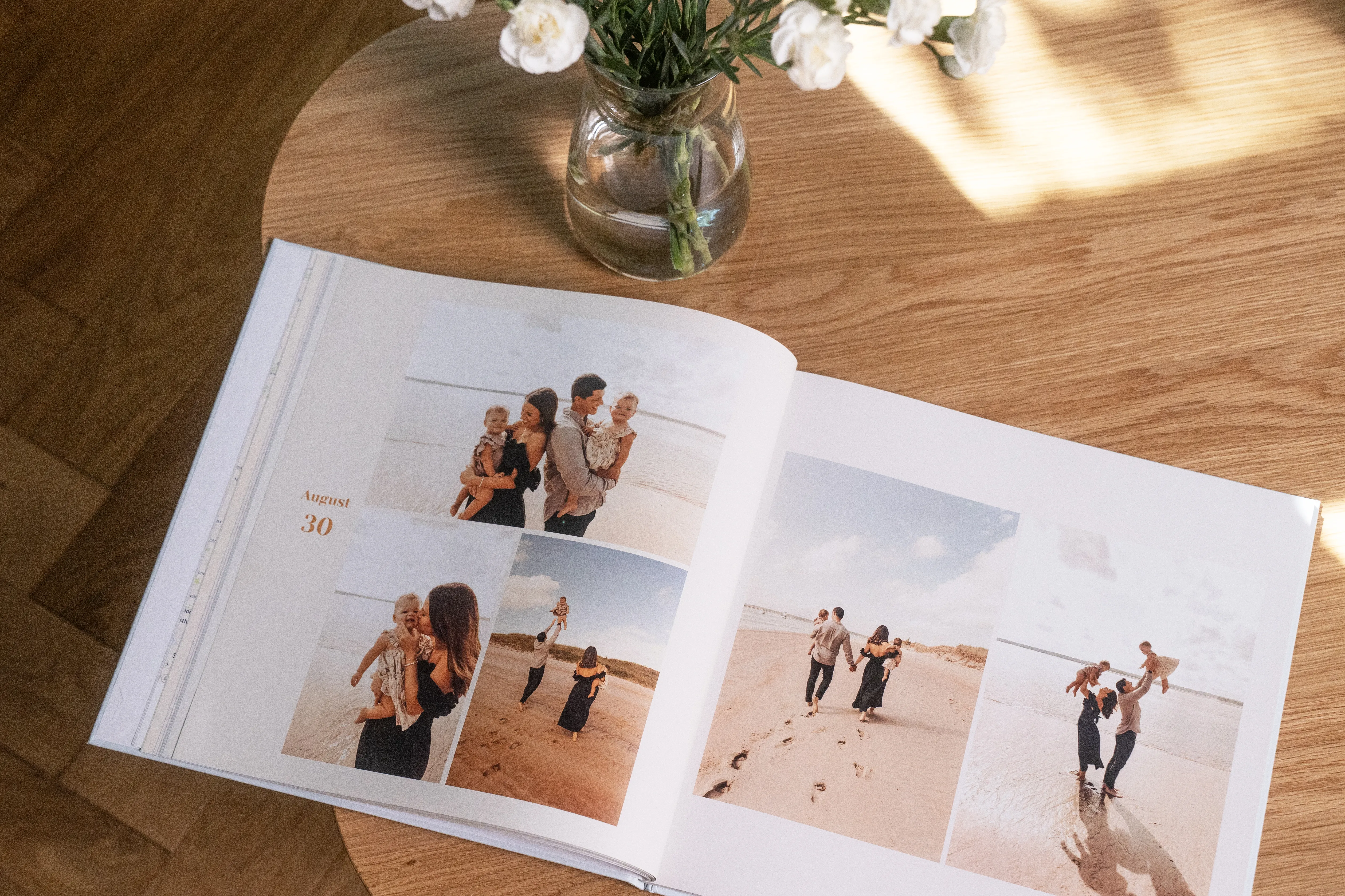 Open photo album on a wooden table, showing family beach photos. A vase with white flowers sits nearby, casting soft shadows.