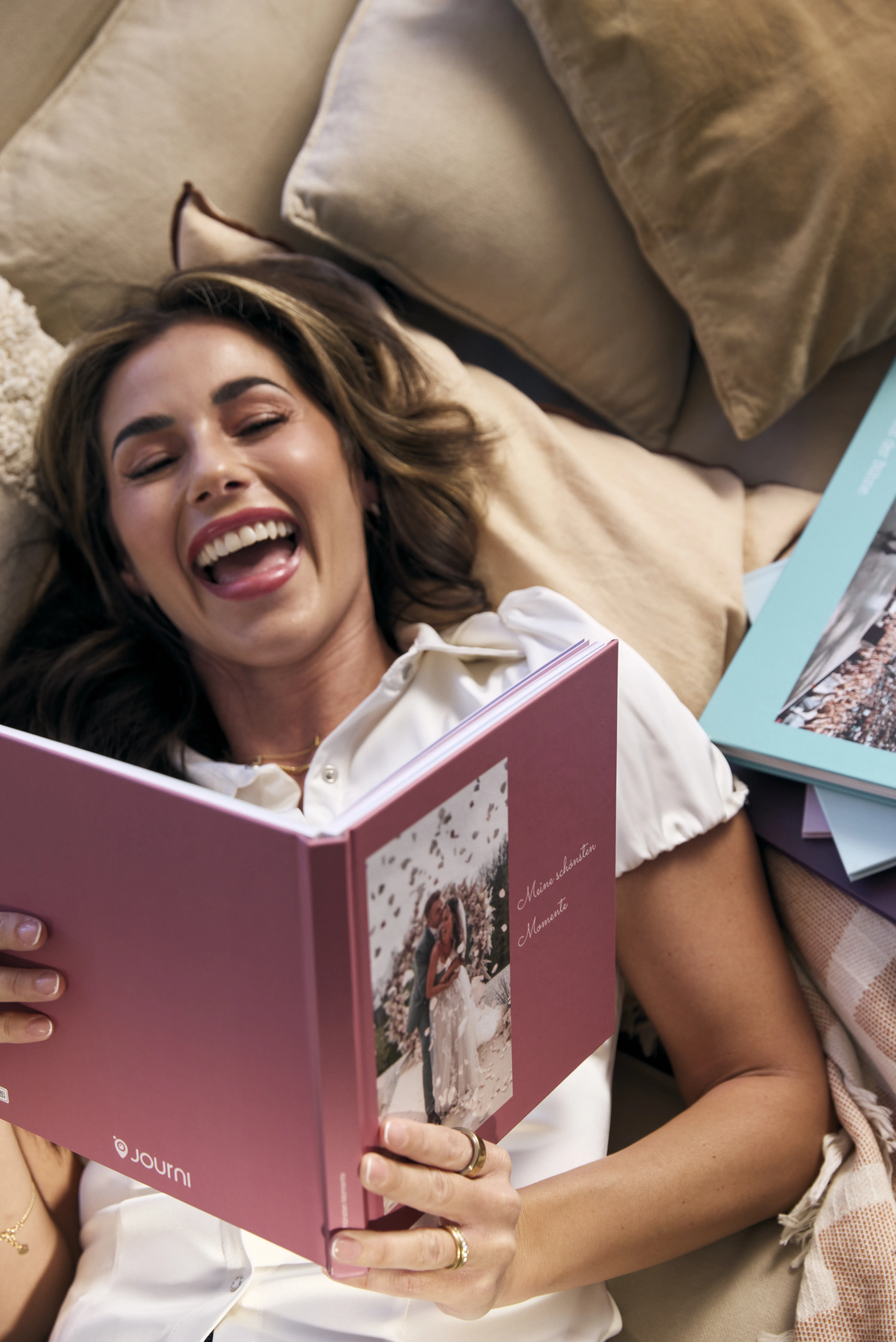 Sarah Engels liegt lachend auf einer Couch und hält ihre Hochzeitsfotobuch in den Händen.