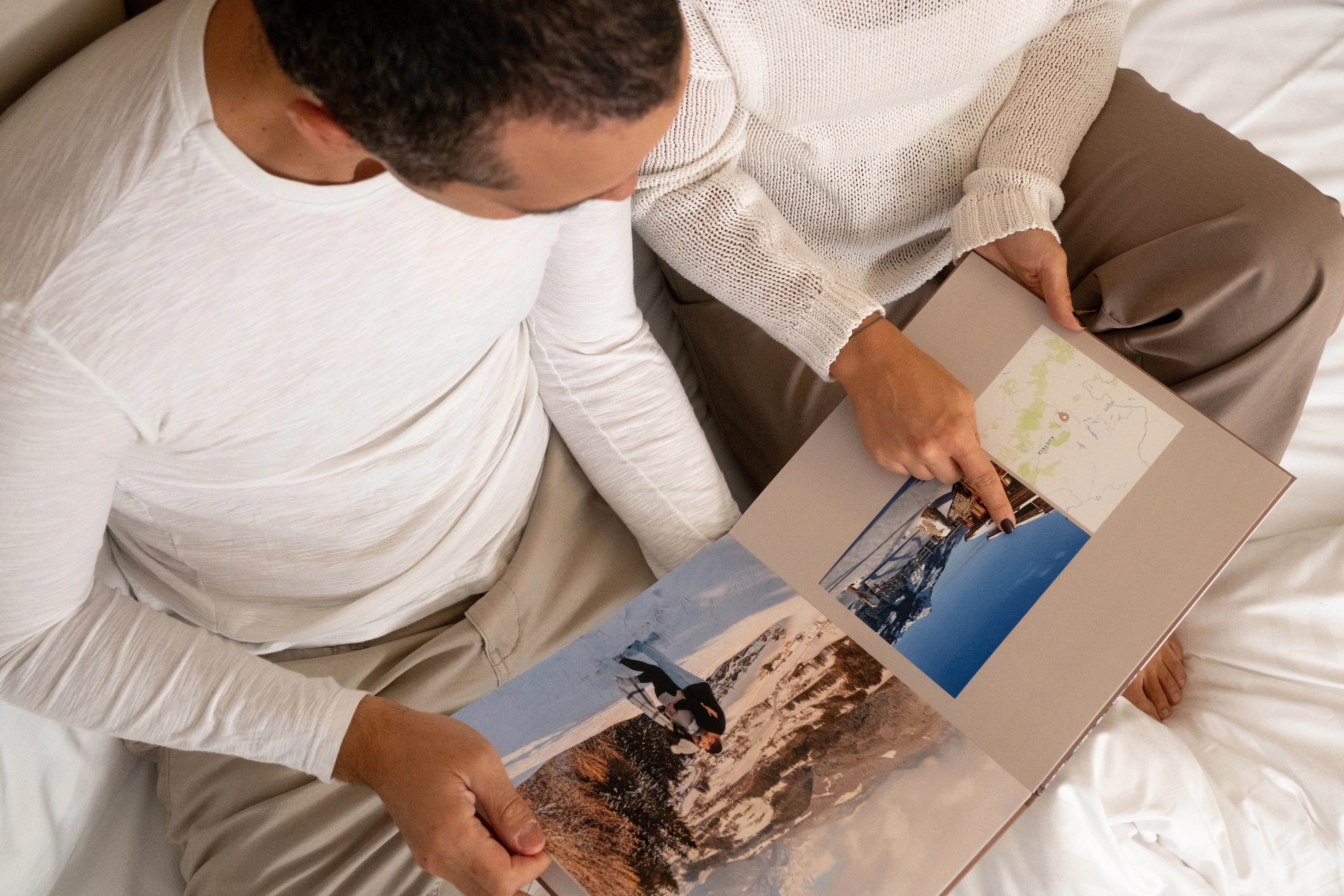 Two people sitting on a bed wearing casual clothing, looking at a photo album with travel photos and a map.