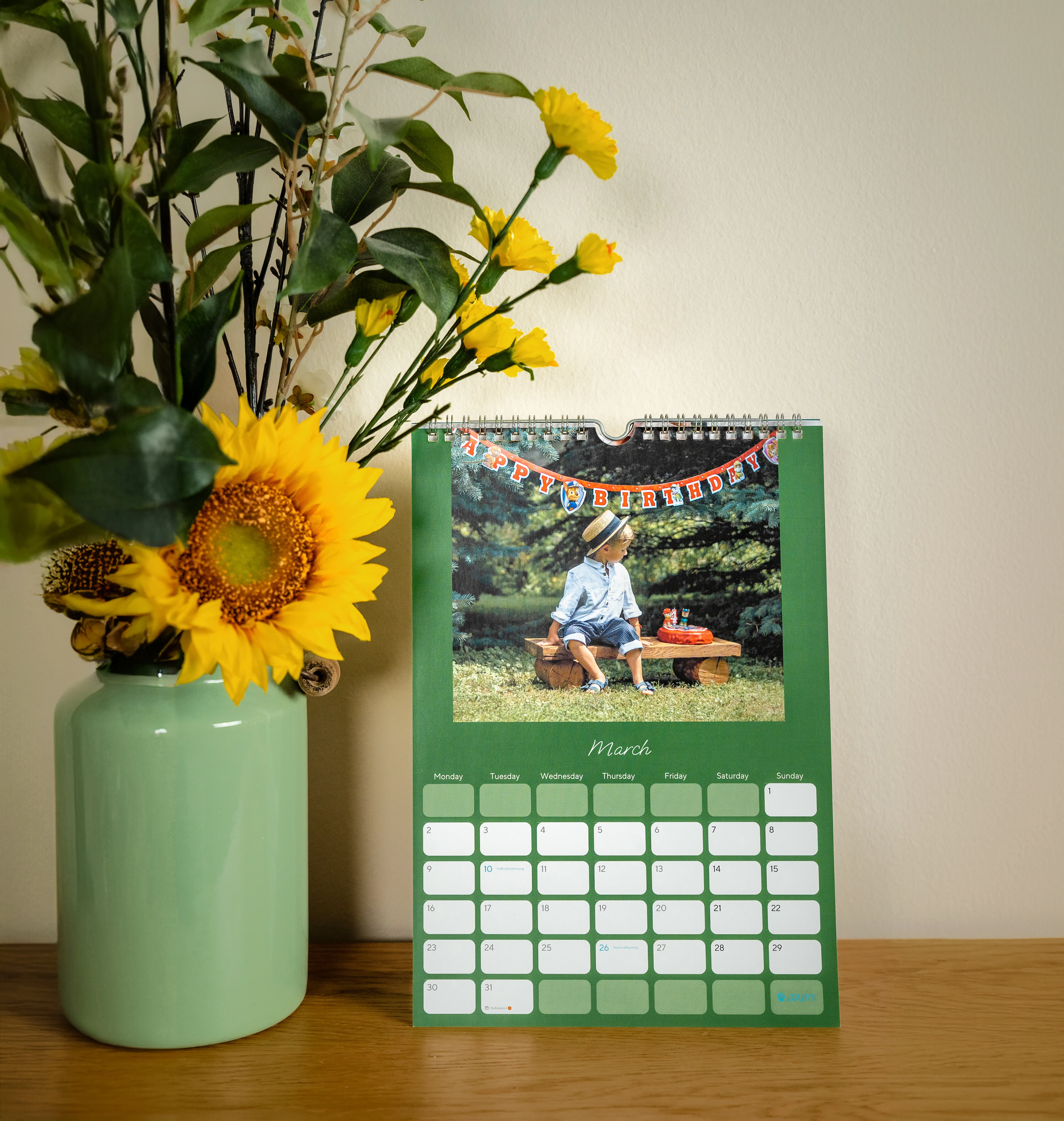 Calendar on a wooden table with a March page showing a photo of a girl in a hat, next to a green vase with sunflowers and greenery.