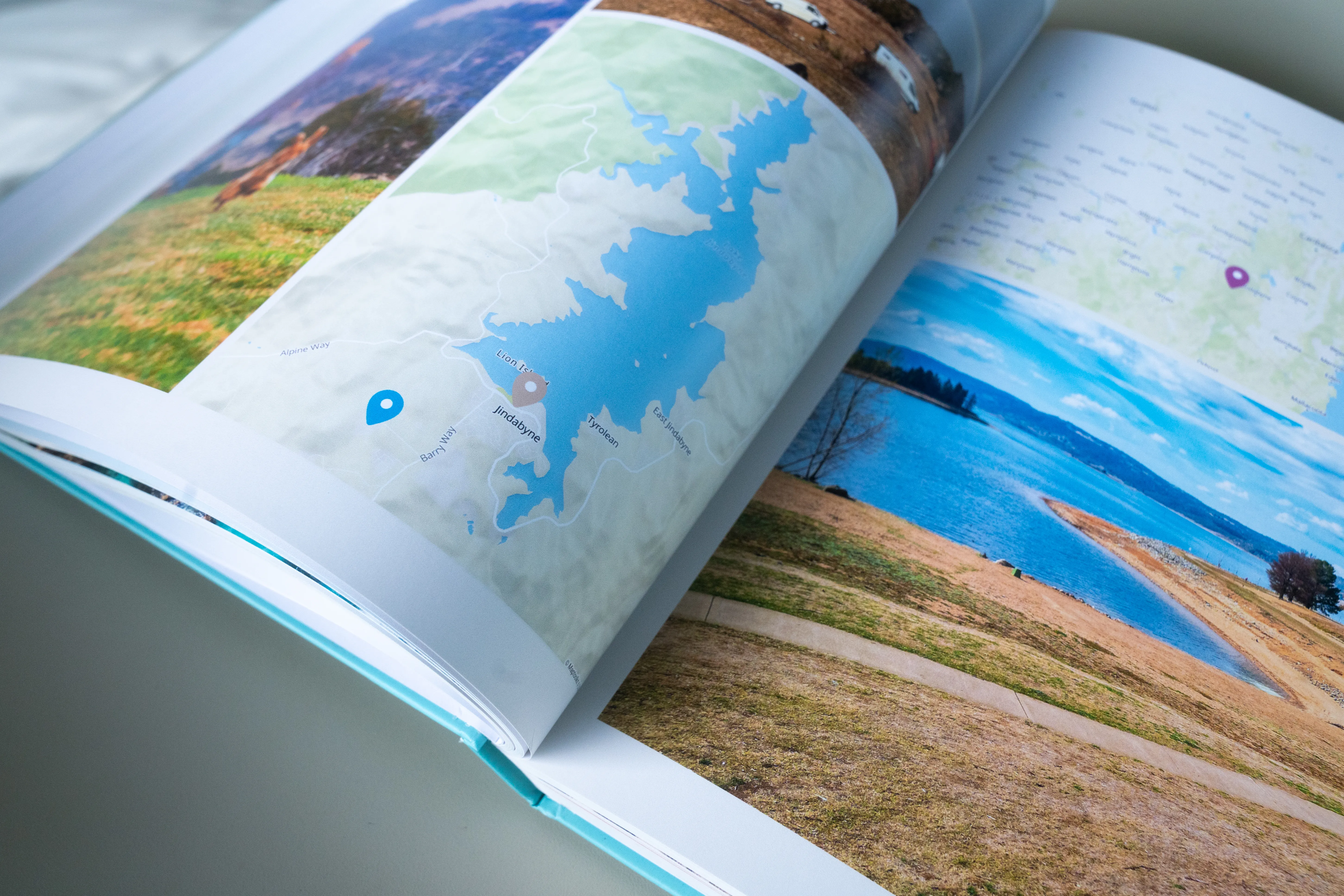 Open travel photo book showing maps and landscape photos, including a blue lake and grassy fields, with a location pin on a map.