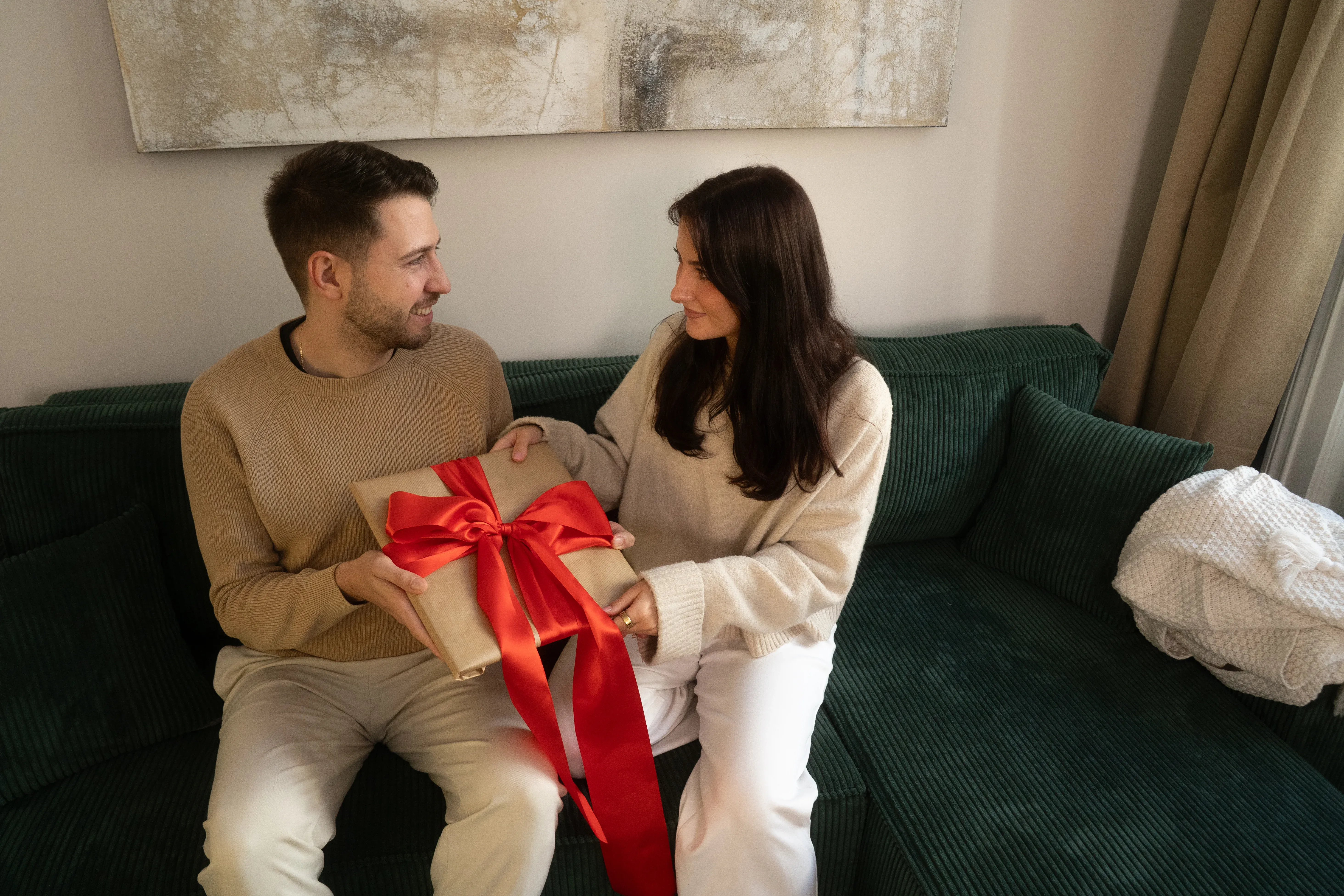 A couple sitting on a green couch, joyfully exchanging a gift wrapped in brown paper with a large red ribbon.