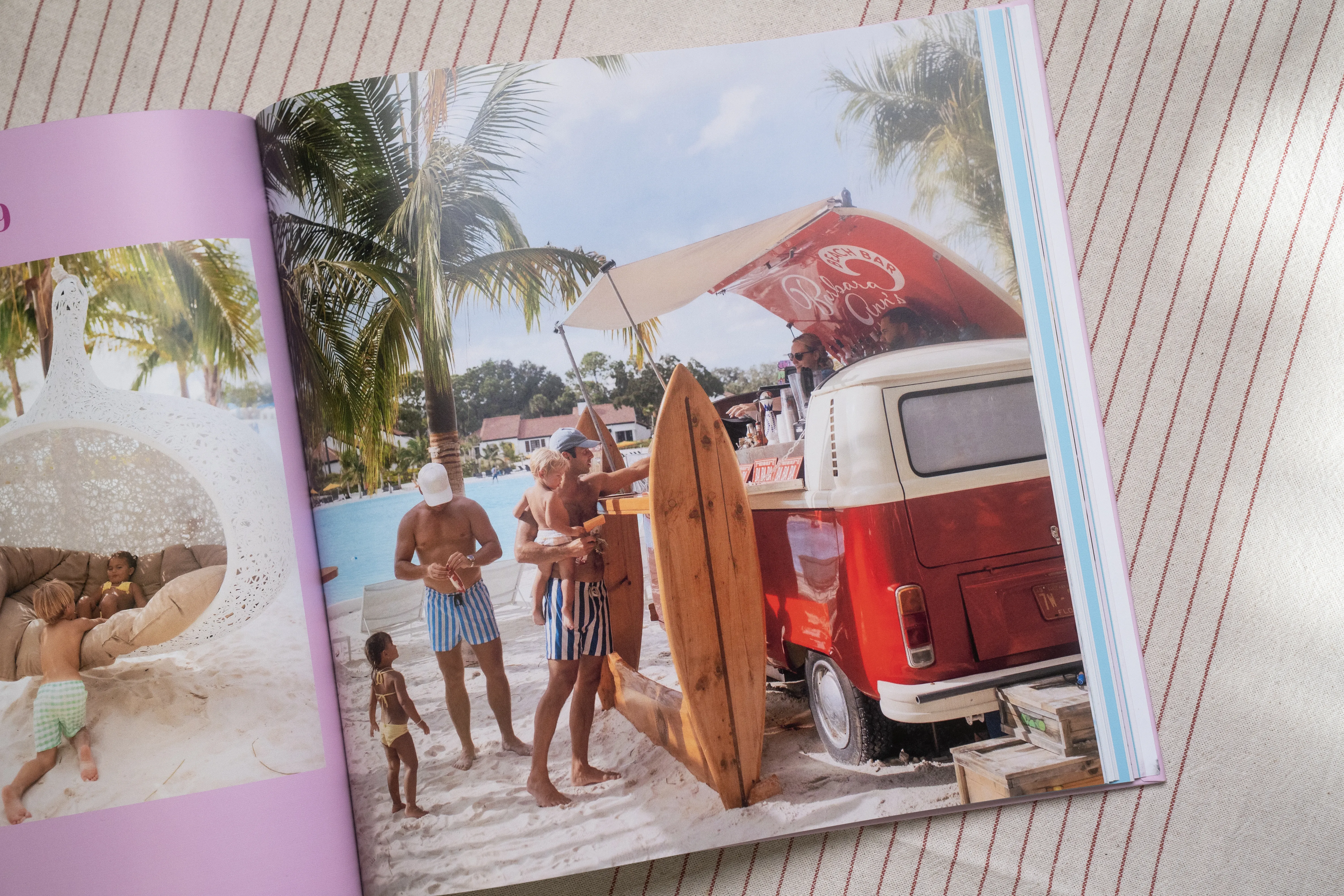 Offenes Buch mit einer Strandszene, wo Leute neben einem Oldtimer-Van, Palmen und einem Surfbrett stehen, vor einem gestreiften Stoffhintergrund.