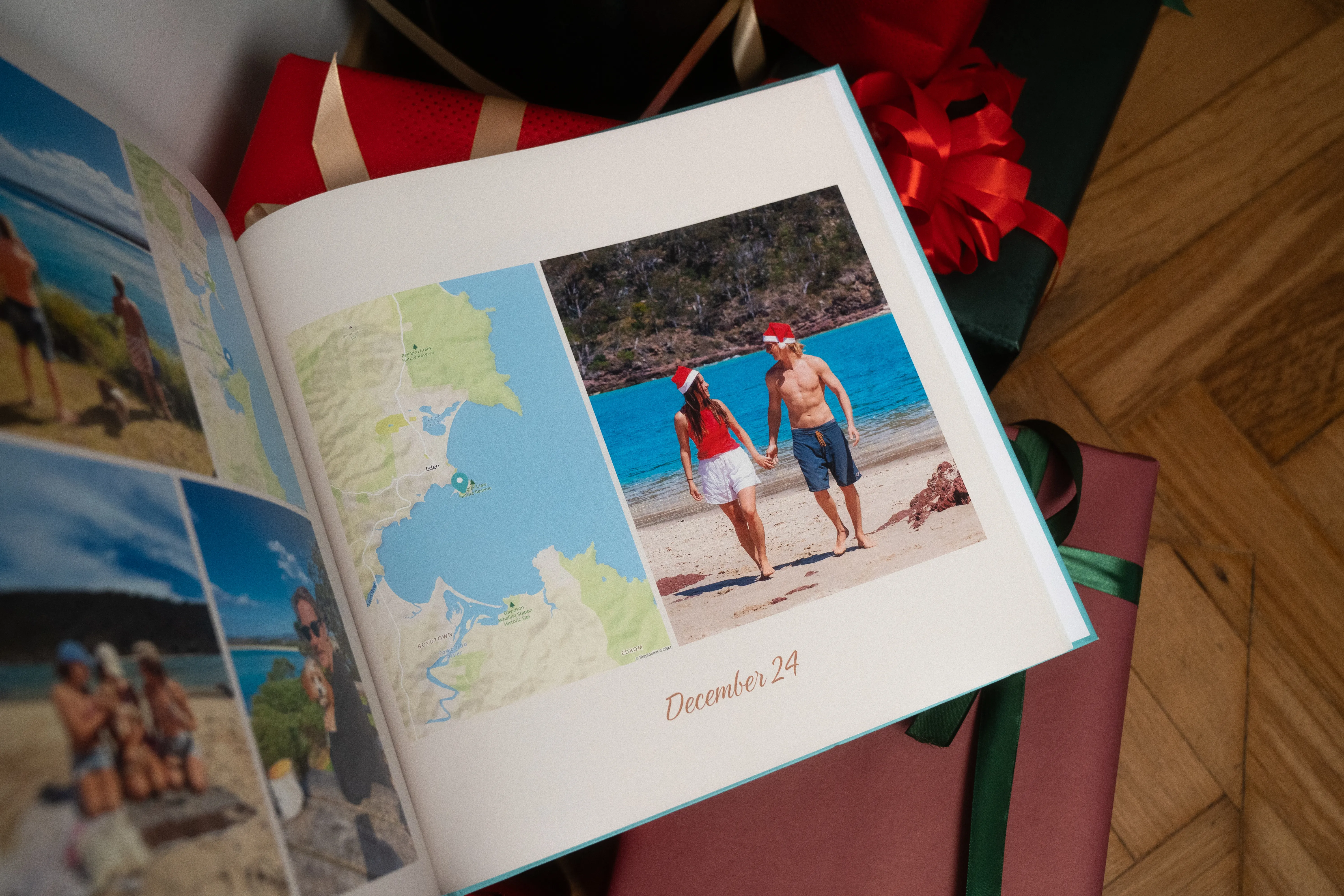 A photo book opened to a page showing a couple in Santa hats walking on a beach, with a map and "December 24" caption, surrounded by wrapped gifts.