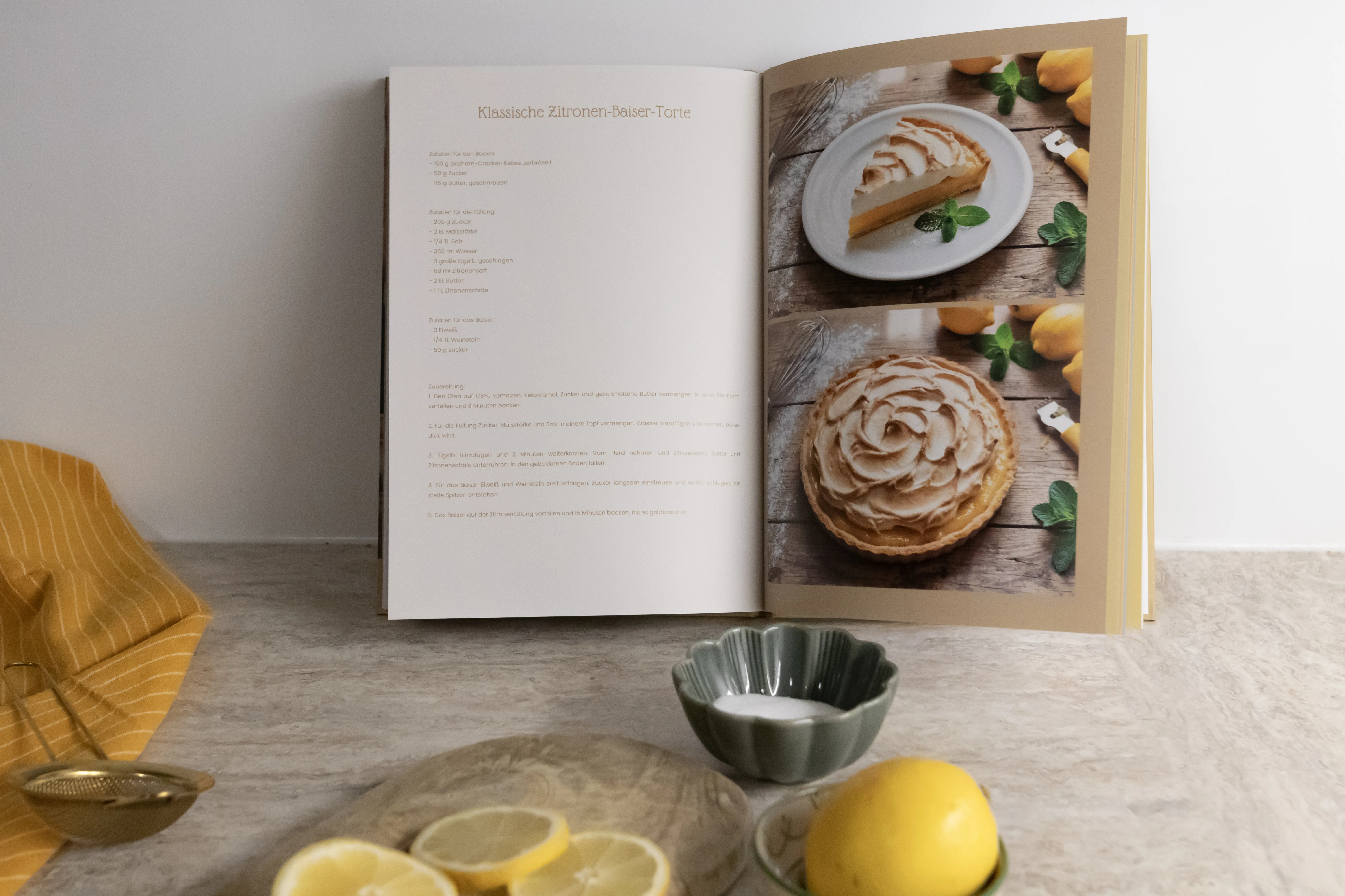 Open cookbook displaying a lemon tart recipe with images; fresh lemons and sliced lemon on a counter nearby.