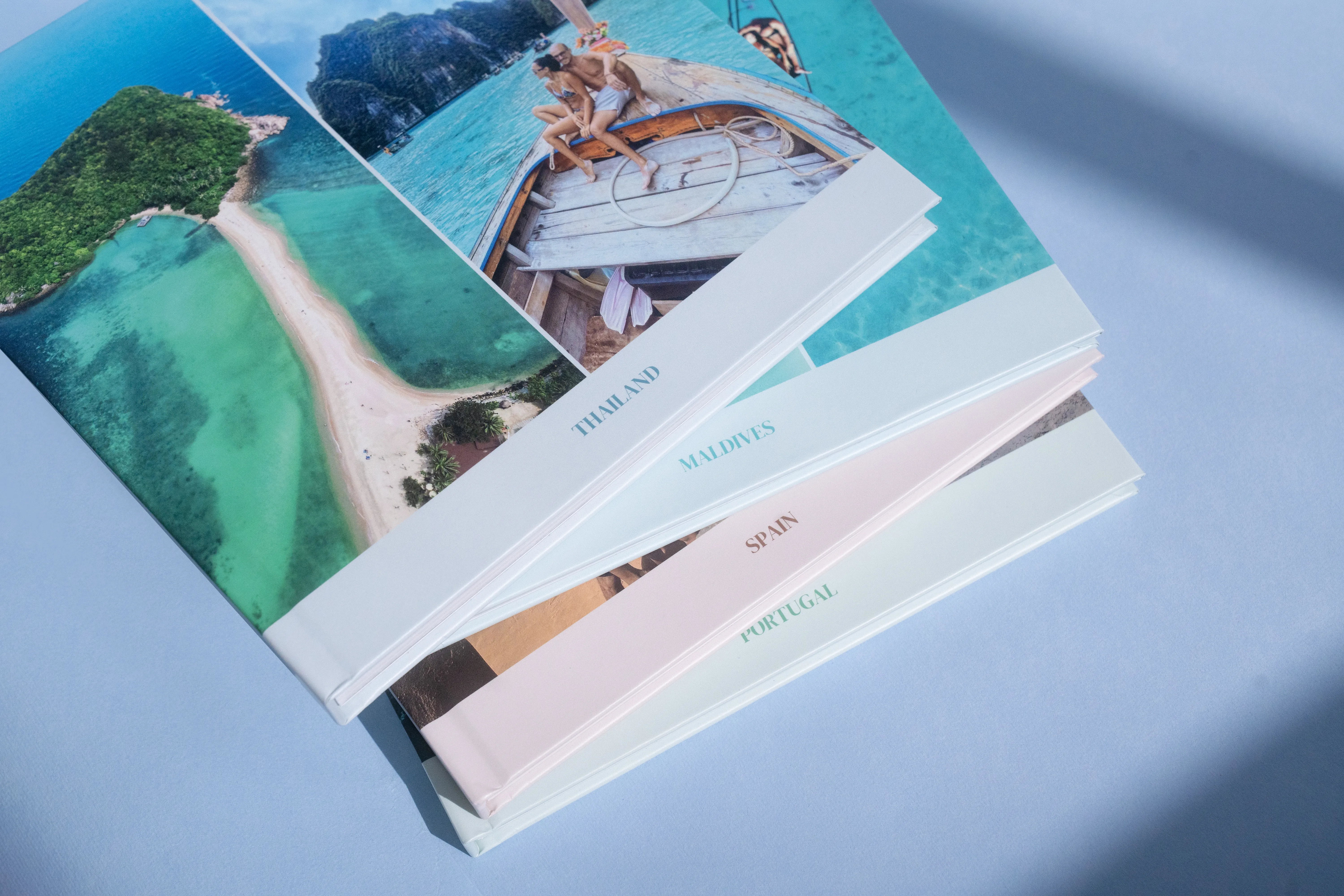 Stack of travel photo books on a light blue surface, featuring images of tropical beaches and destinations like Thailand and Portugal.
