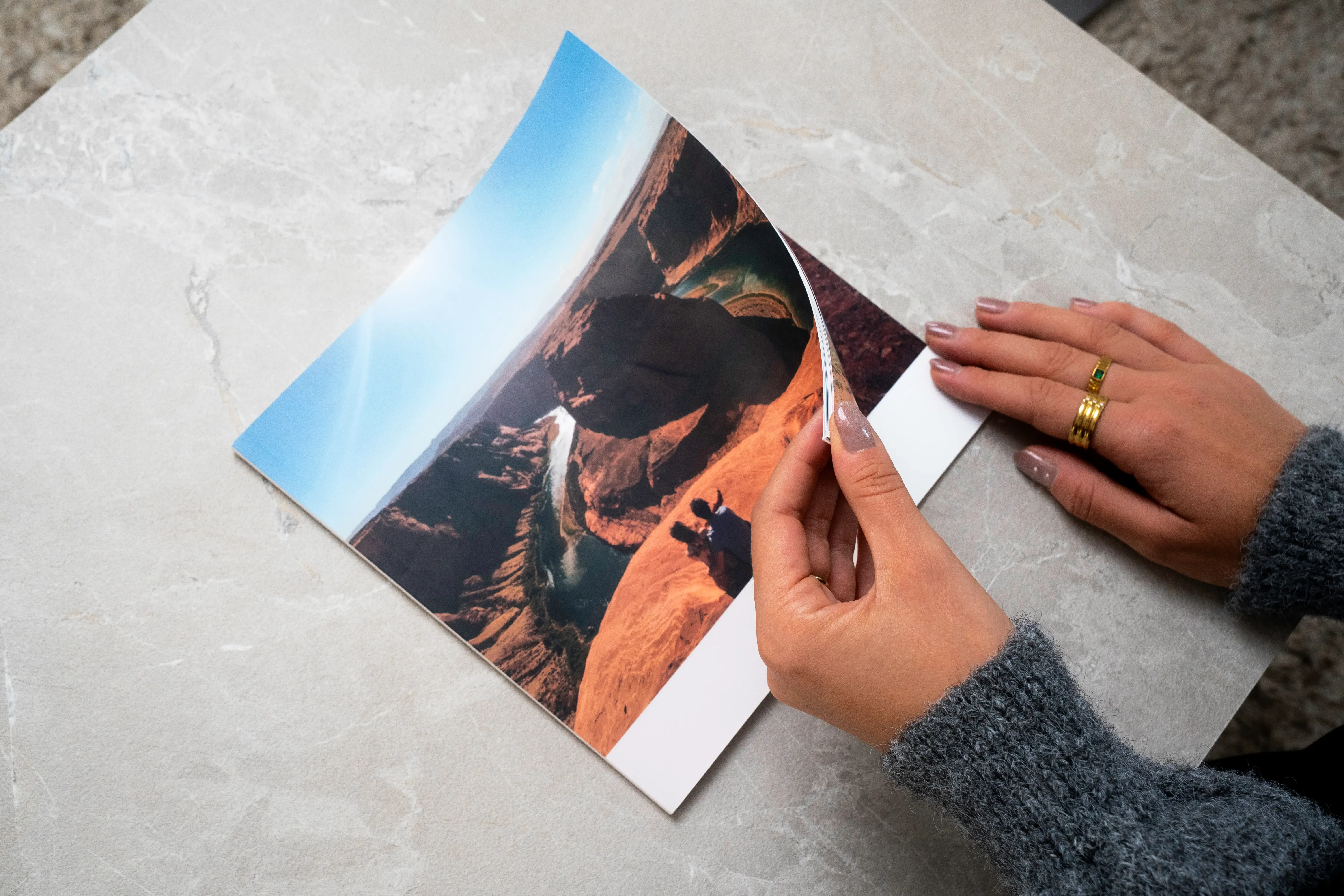 Hands flipping through a softcover photo book with an image of a scenic canyon. The surface is a light-colored stone table.