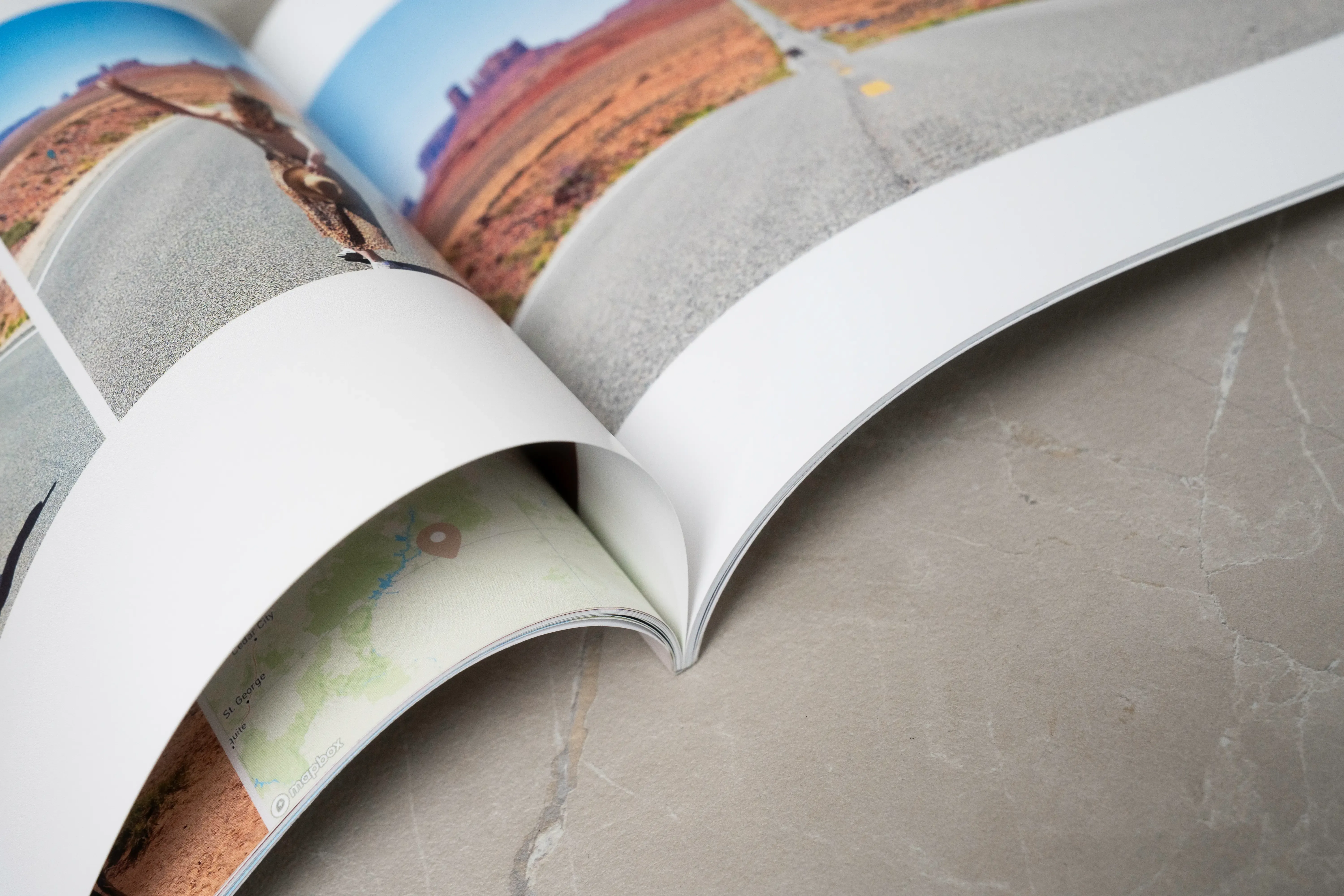 Open softcover photo book on a marble surface, featuring landscape photos and partially visible pages.