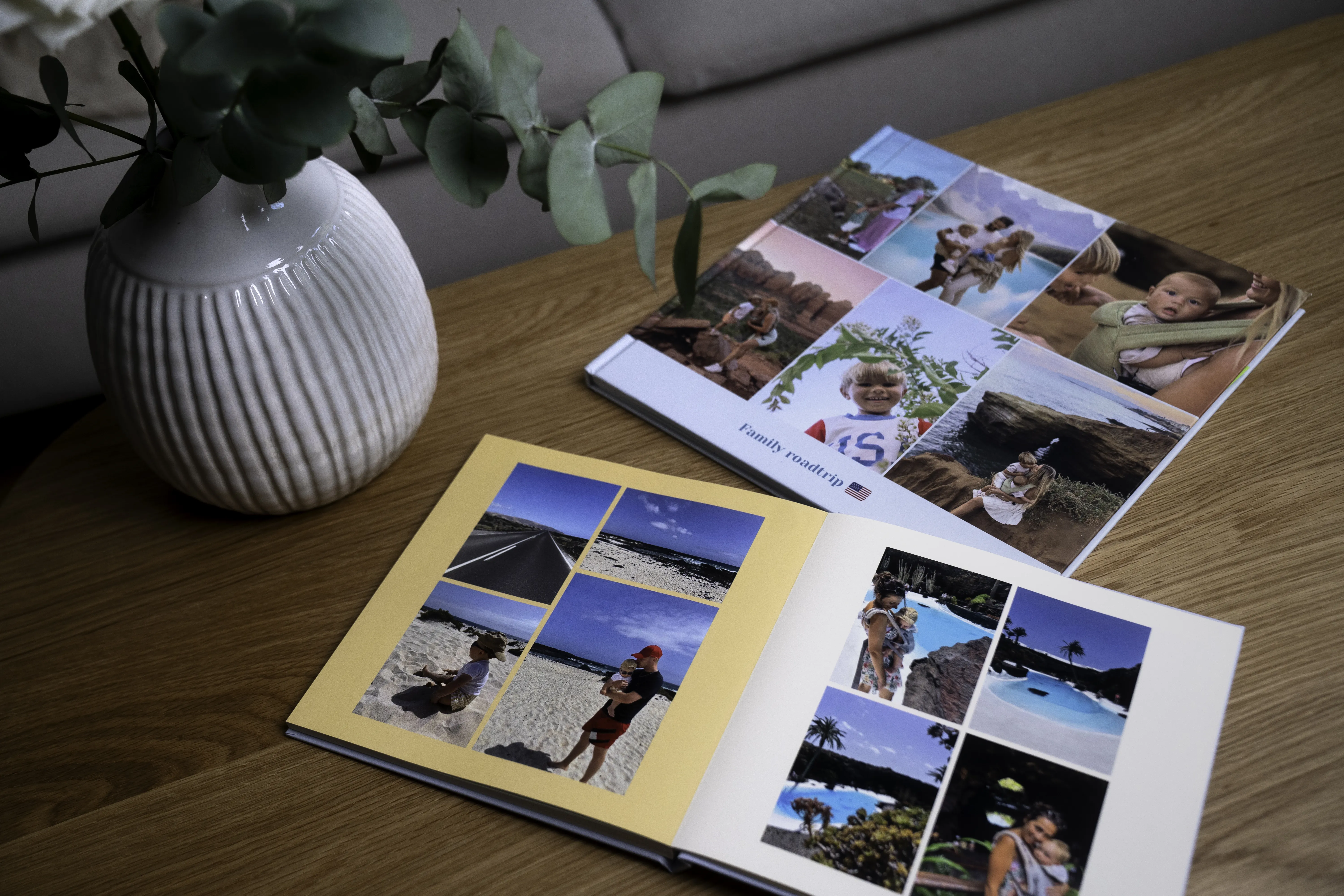 Two open photo albums with vacation pictures on a wooden table next to a white vase with green foliage.