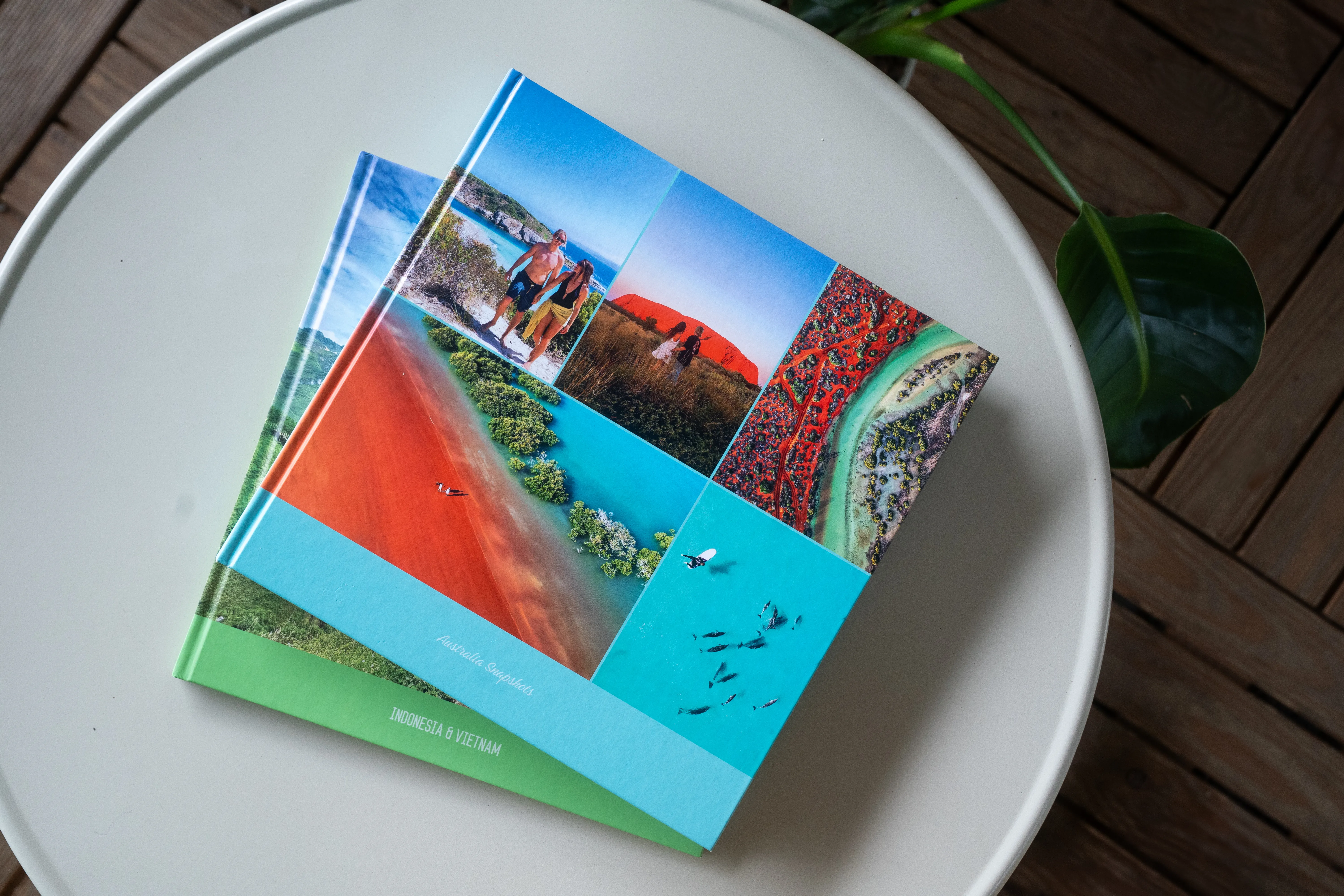 Ein farbenfrohes Fotobuch auf einem runden Tisch, dessen Einband mit lebendigen Natur- und Landschaftsbildern verziert ist. Eine Pflanze ist teilweise zu sehen.