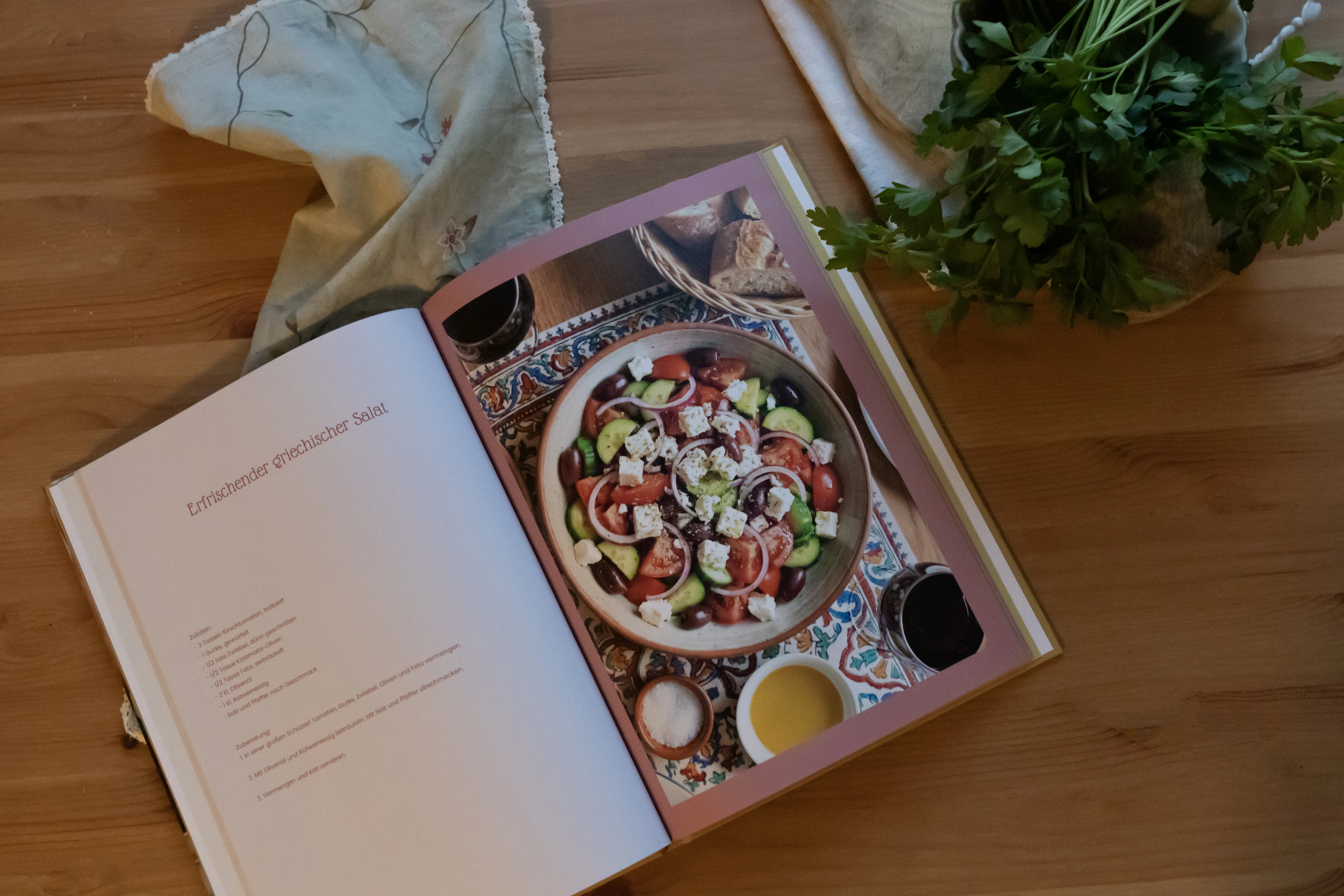 Offenes Kochbuch auf einem Holztisch mit einem Rezept für griechischen Salat, daneben frische Kräuter und ein Tuch.