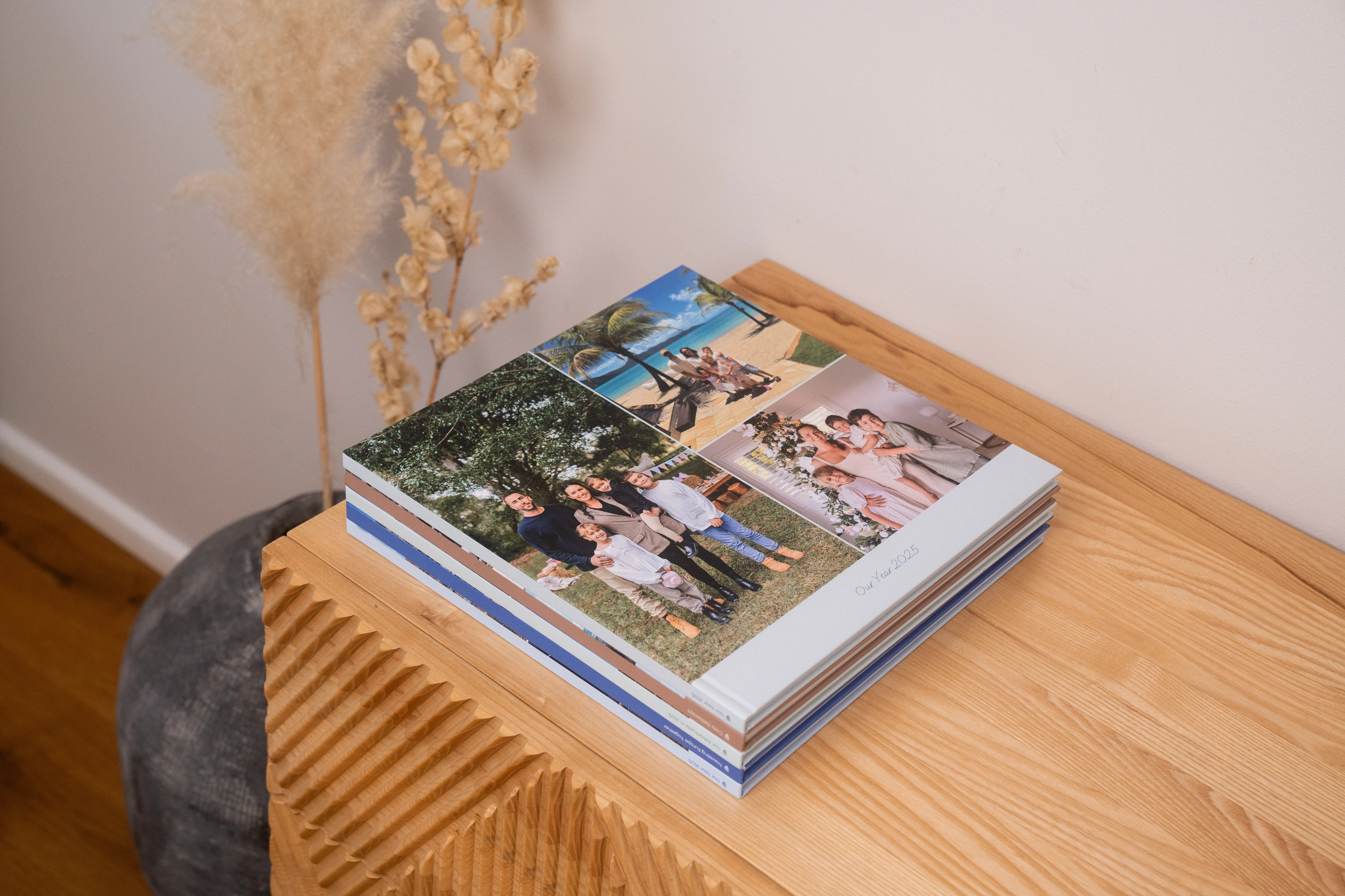 A stack of photo albums with family pictures on a wooden table, next to dried plants in a vase.
