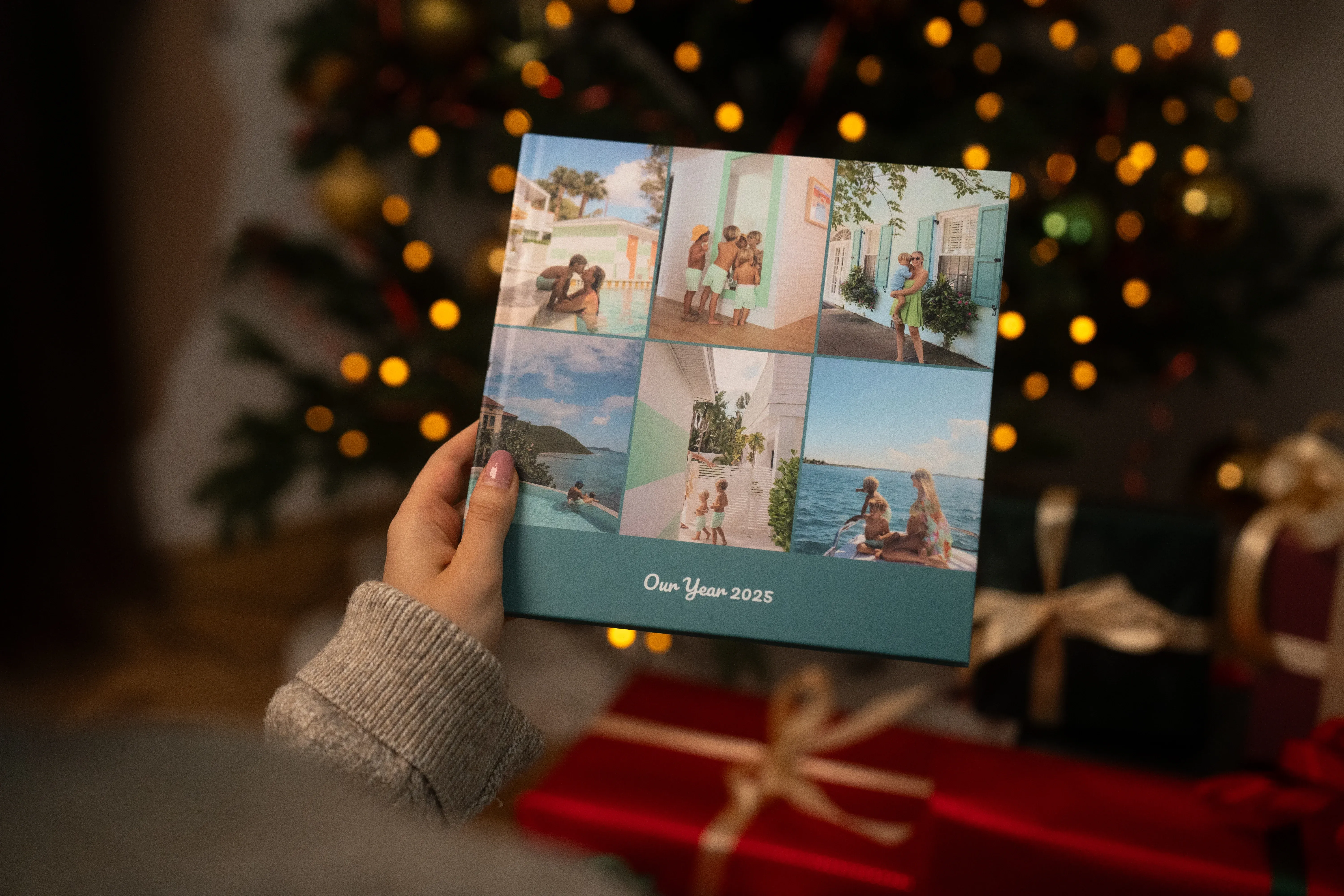 Eine Person hält ein Fotobuch mit dem Titel „Unser Jahr 2023” vor einem geschmückten Weihnachtsbaum und verpackten Geschenken in der Hand.